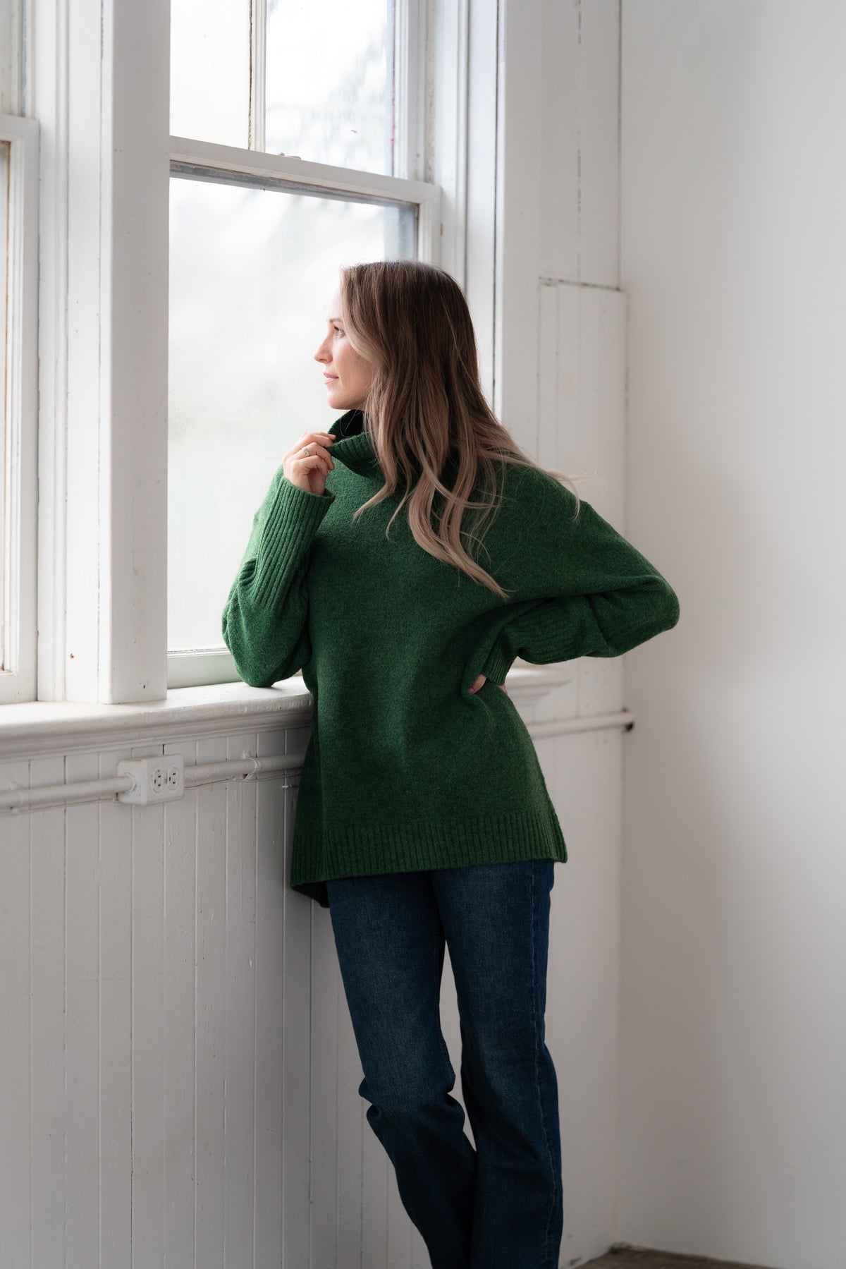 Woman wearing the Echoverde Dog Walker forest green chunky cowl neck sweater with dark jeans, pulling the cowl up over her chin