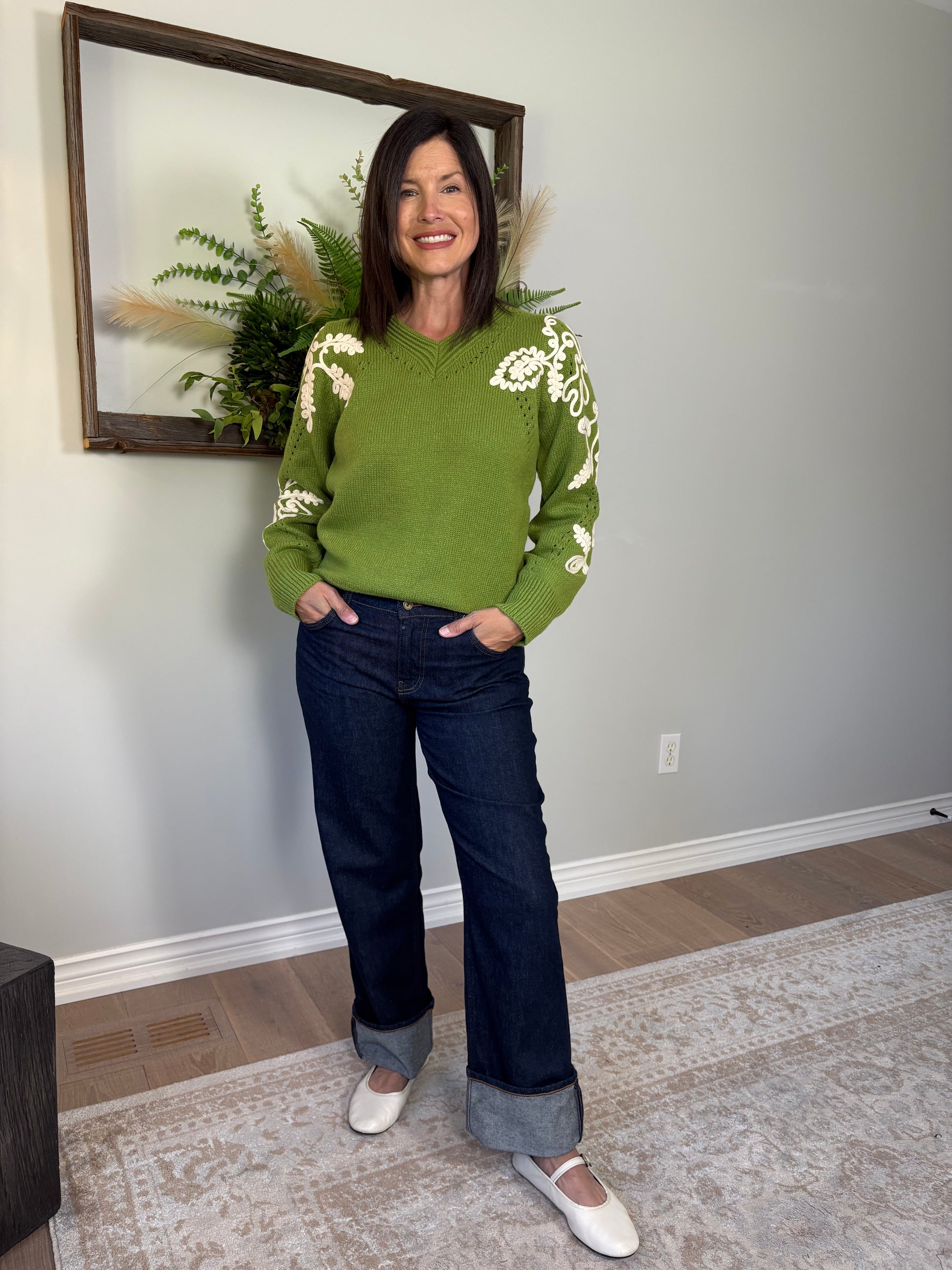 Woman wearing a green sweater with white patterns and dark jeans indoors.