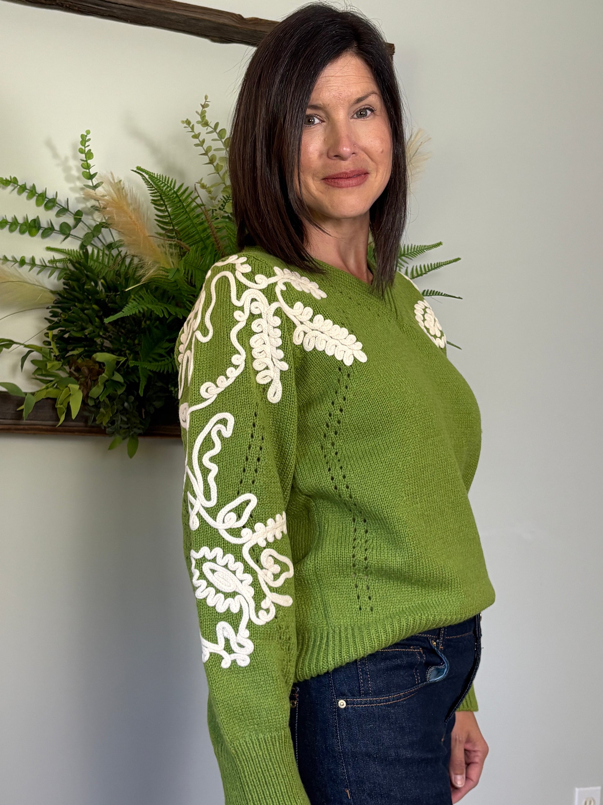 Woman wearing a green sweater with white floral patterns, standing against a neutral wall with decorative plants.
