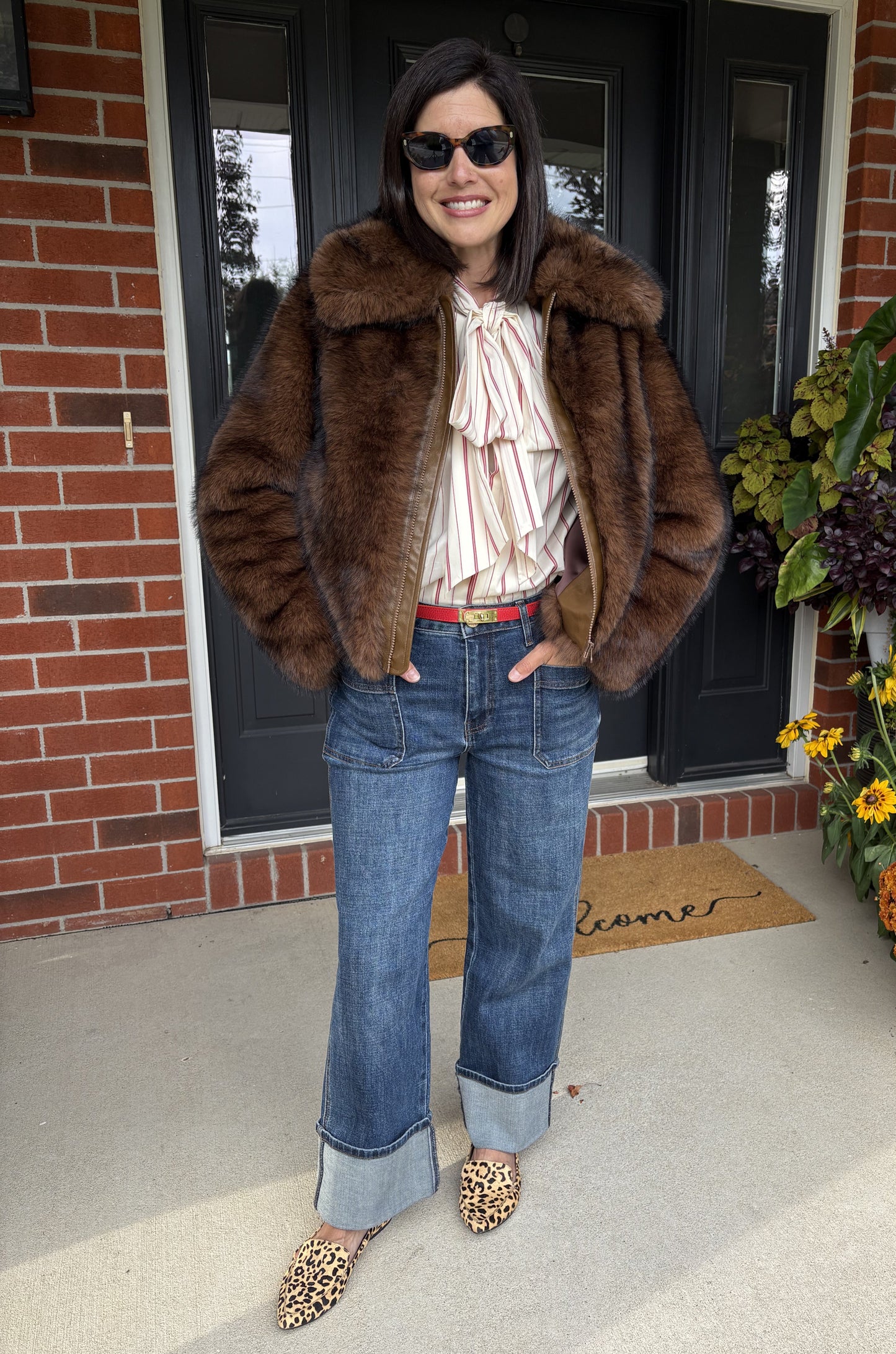 Woman in a fur coat and jeans standing on a porch with flowers and a welcome mat.