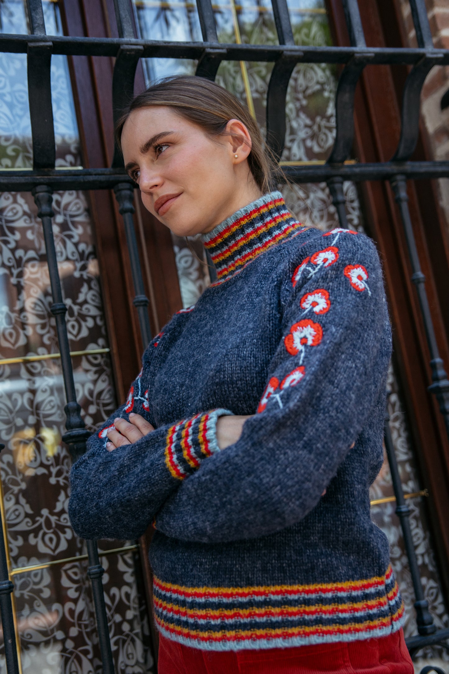 Woman wearing a blue sweater with colorful patterns standing in front of a decorative metal gate.