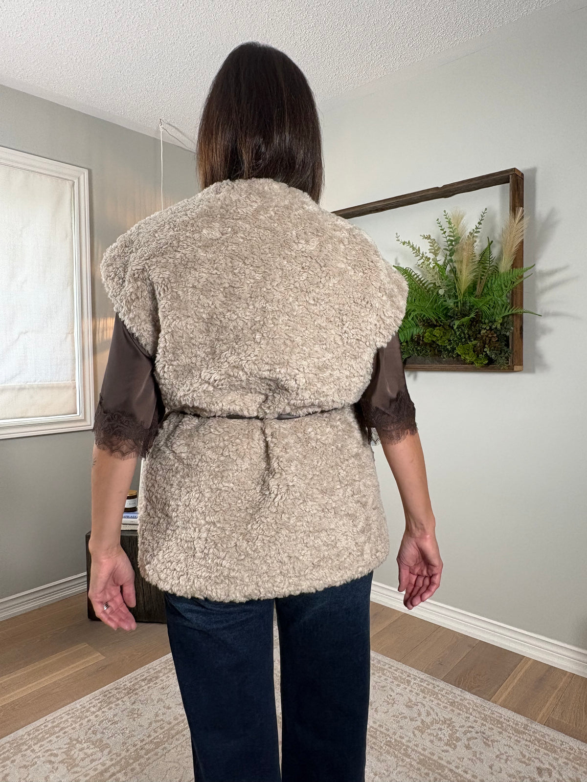 A woman viewed from behind wearing a beige textured faux-shearling belted vest over sheer dark brown sleeves, paired with dark blue jeans, standing in a home interior.