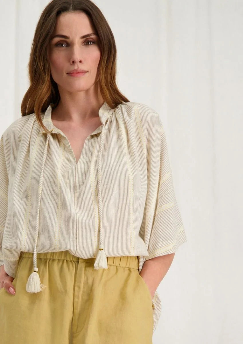 Introducing the Avril Blouse in Antique White by Circle of Trust. Made from soft 100% cotton, it's comfy and breathable for everyday wear. The loose fit and vertical stripe pattern give it a relaxed yet stylish vibe. With a V-neck and delicate strings, it's effortlessly charming. Plus, it has drop shoulders and wide sleeves for a comfy fit. Finished with tassel ties and subtle ochre accents, it's a versatile piece for any occasion. Pair it with denim shorts or a skirt for a casual look. 