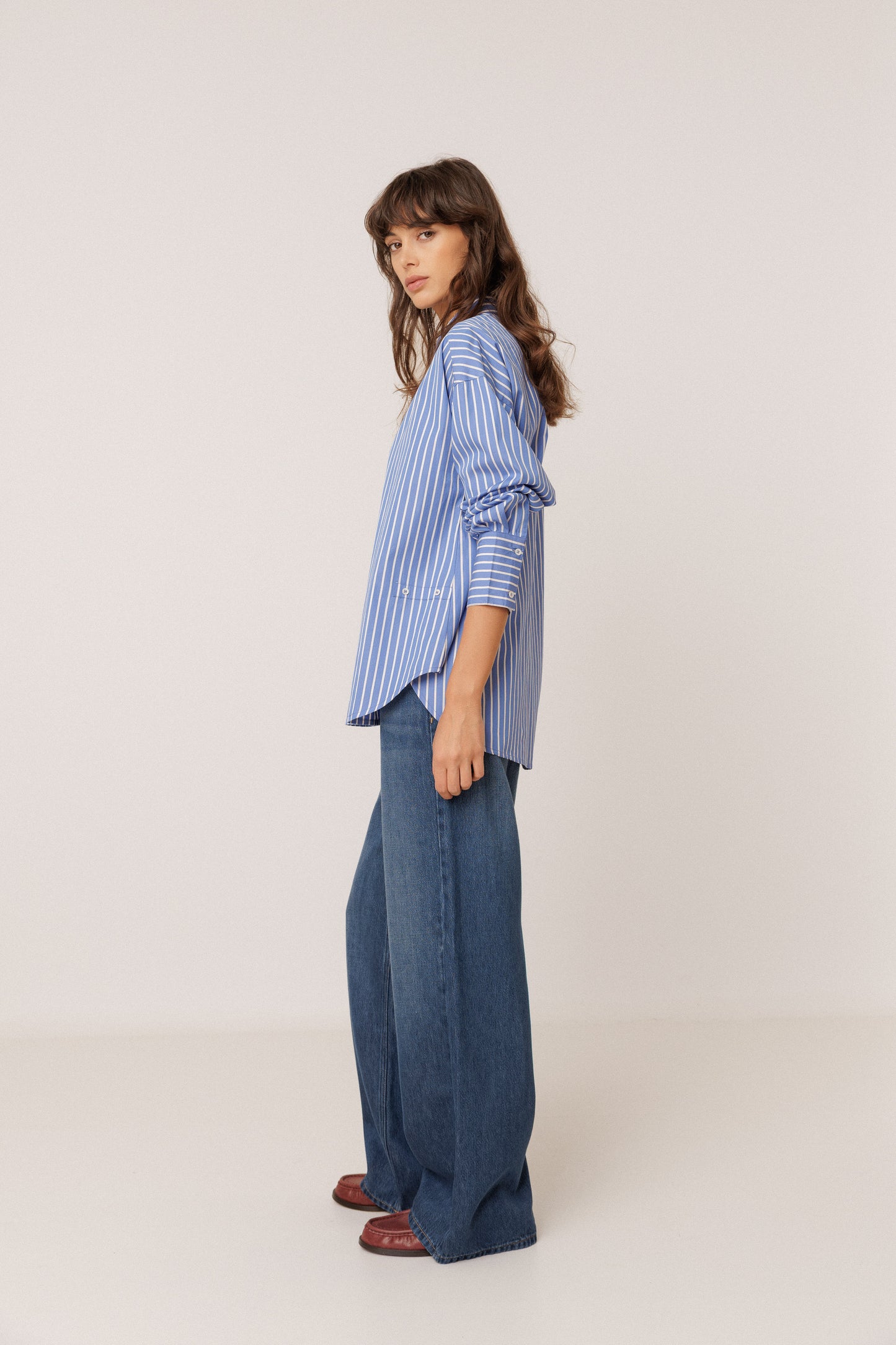 A woman stands in profile wearing a blue and white vertically striped button-up shirt with ruffled sleeves, wide-leg blue jeans, and brown shoes, photographed against a plain light beige studio background.
