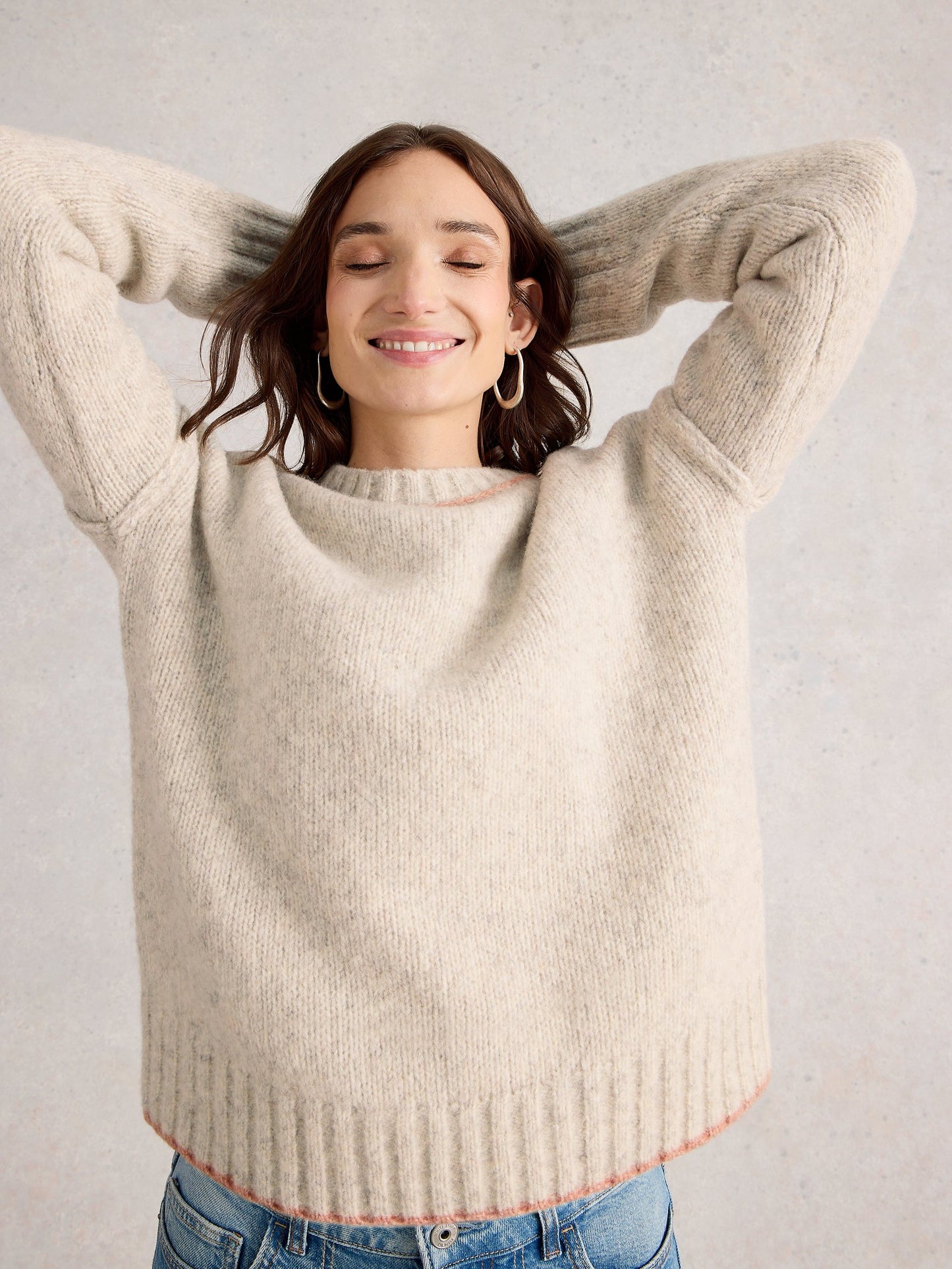 Woman wearing a beige sweater with her hands behind her head against a plain background