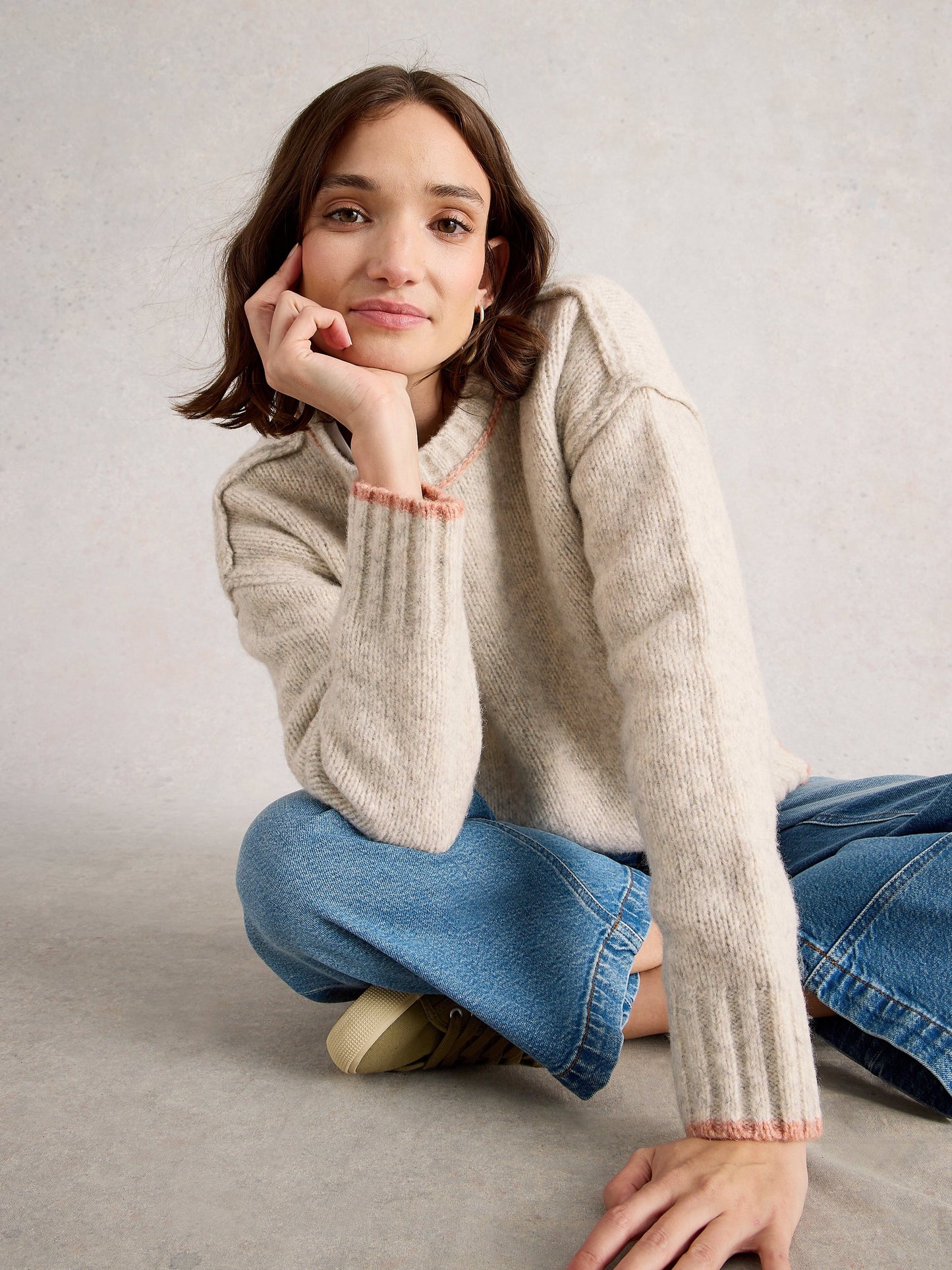 Woman wearing a beige sweater and blue jeans sitting on the floor against a plain background