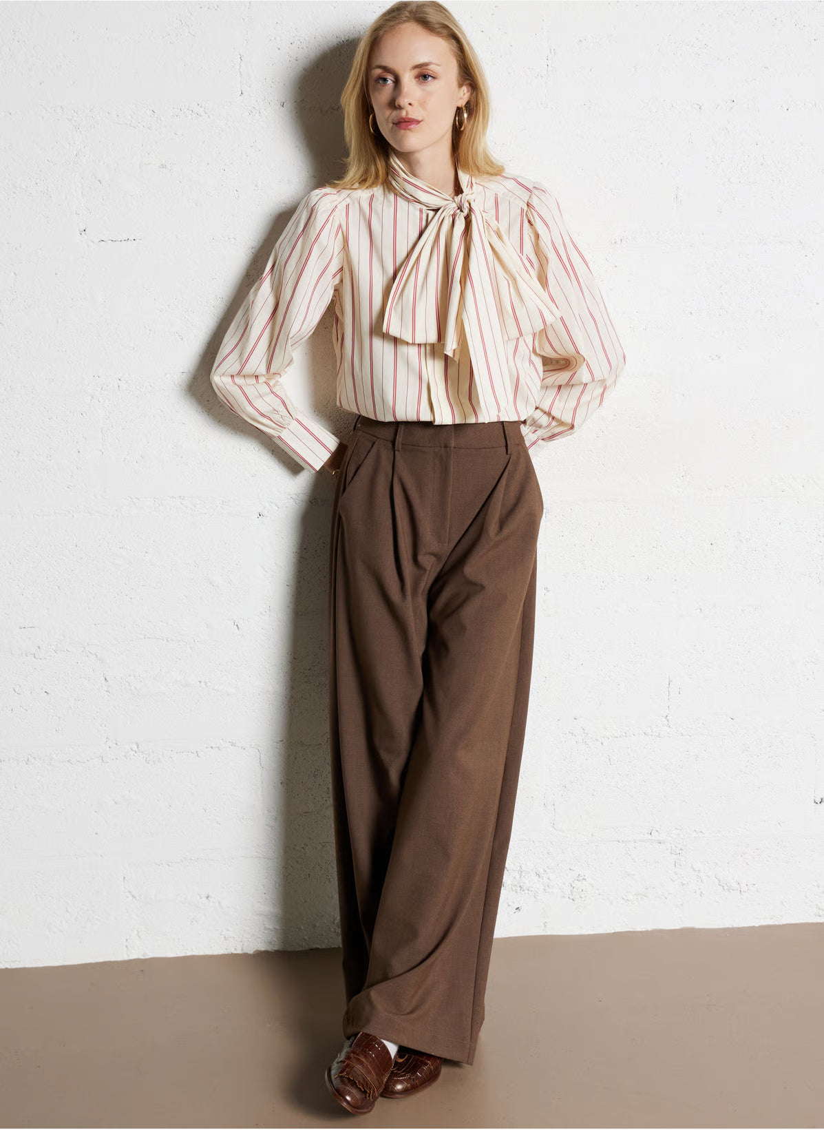 Woman wearing a striped blouse with a bow and brown pants against a white brick wall.