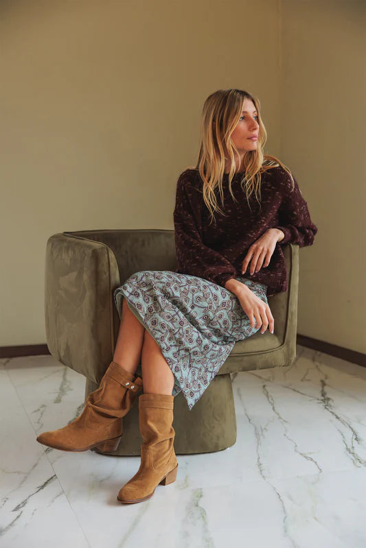 Woman sitting on a green chair wearing a brown sweater, floral skirt, and tan boots.