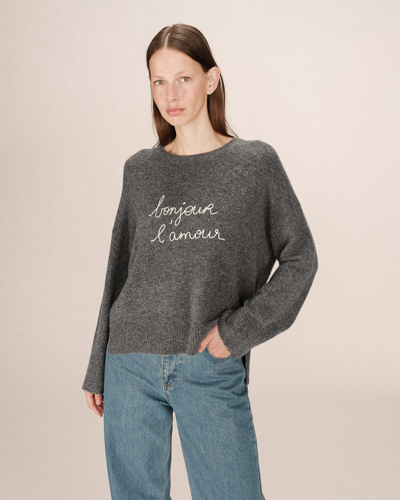 Woman wearing a gray sweater with 'bonjour l'amour' text and blue jeans on a beige background