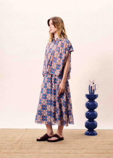 Woman wearing a patterned dress standing next to a blue vase on a beige surface.