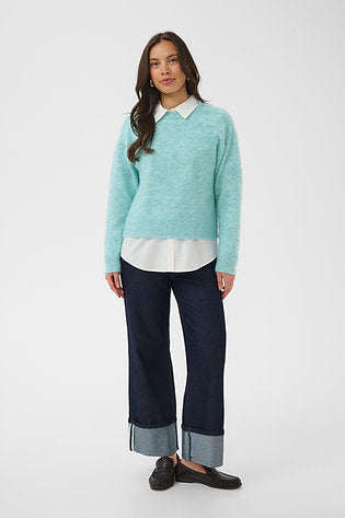 A woman modeling a light aqua knit sweater over a white collared shirt, paired with dark blue cropped jeans and black loafers, against a plain light gray studio background.