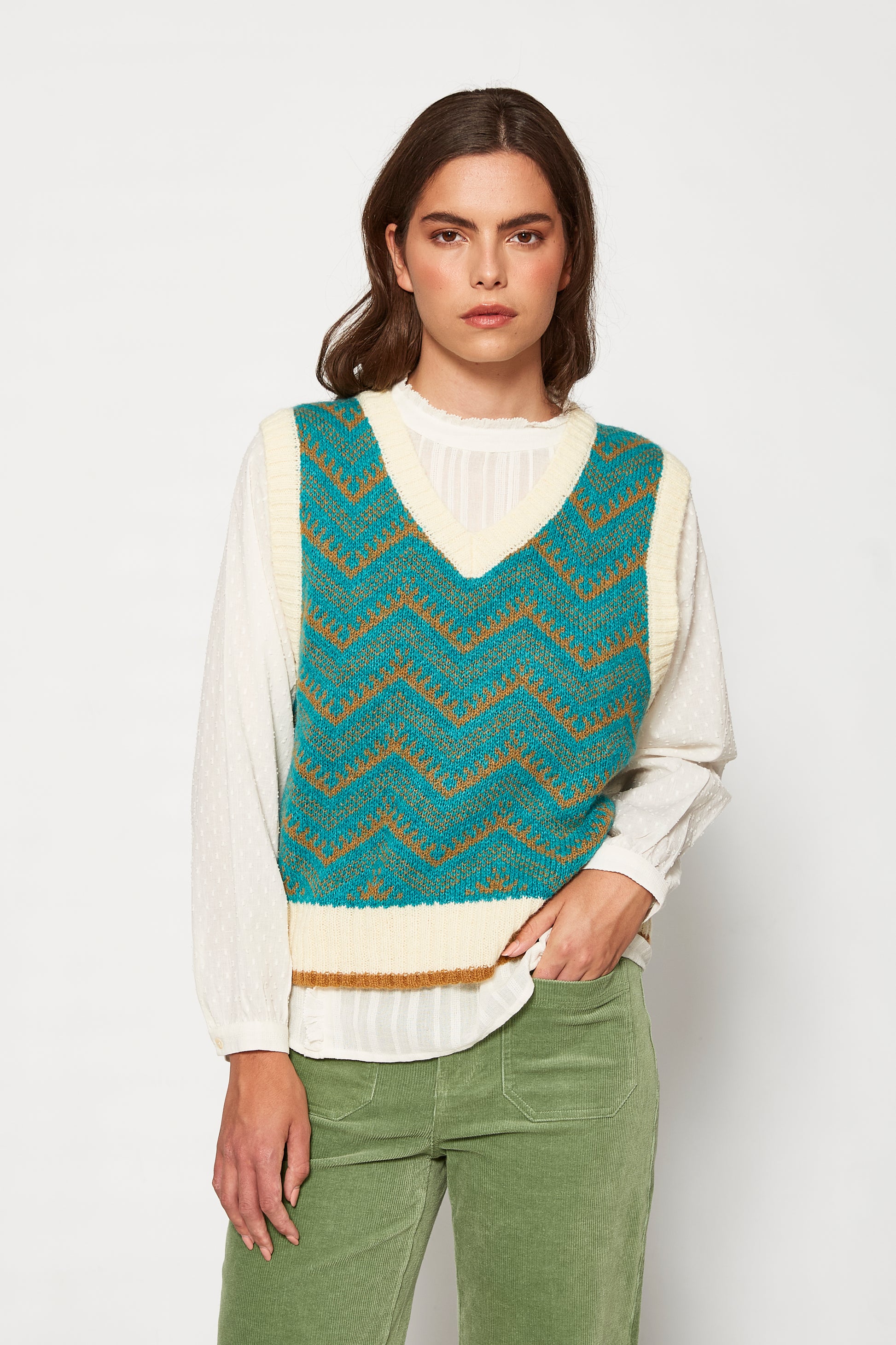 Woman wearing a teal and brown patterned vest over a white shirt with green pants on a white background