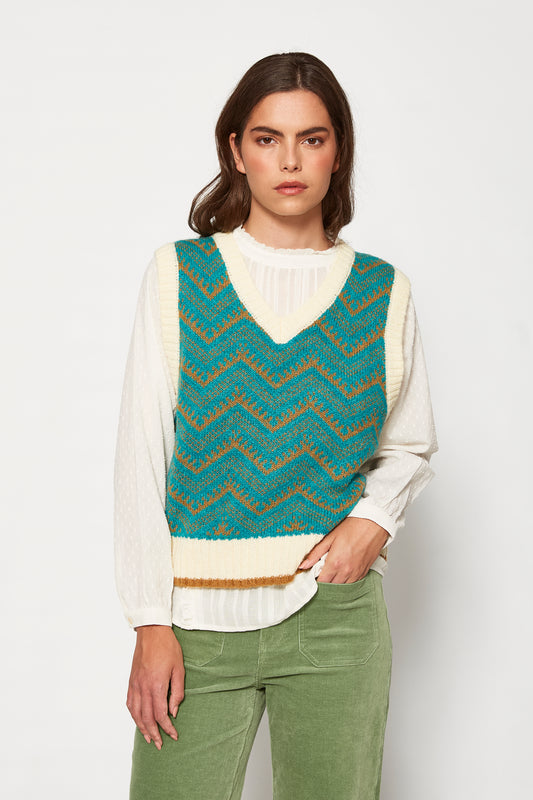 Woman wearing a teal and brown patterned vest over a white shirt with green pants on a white background