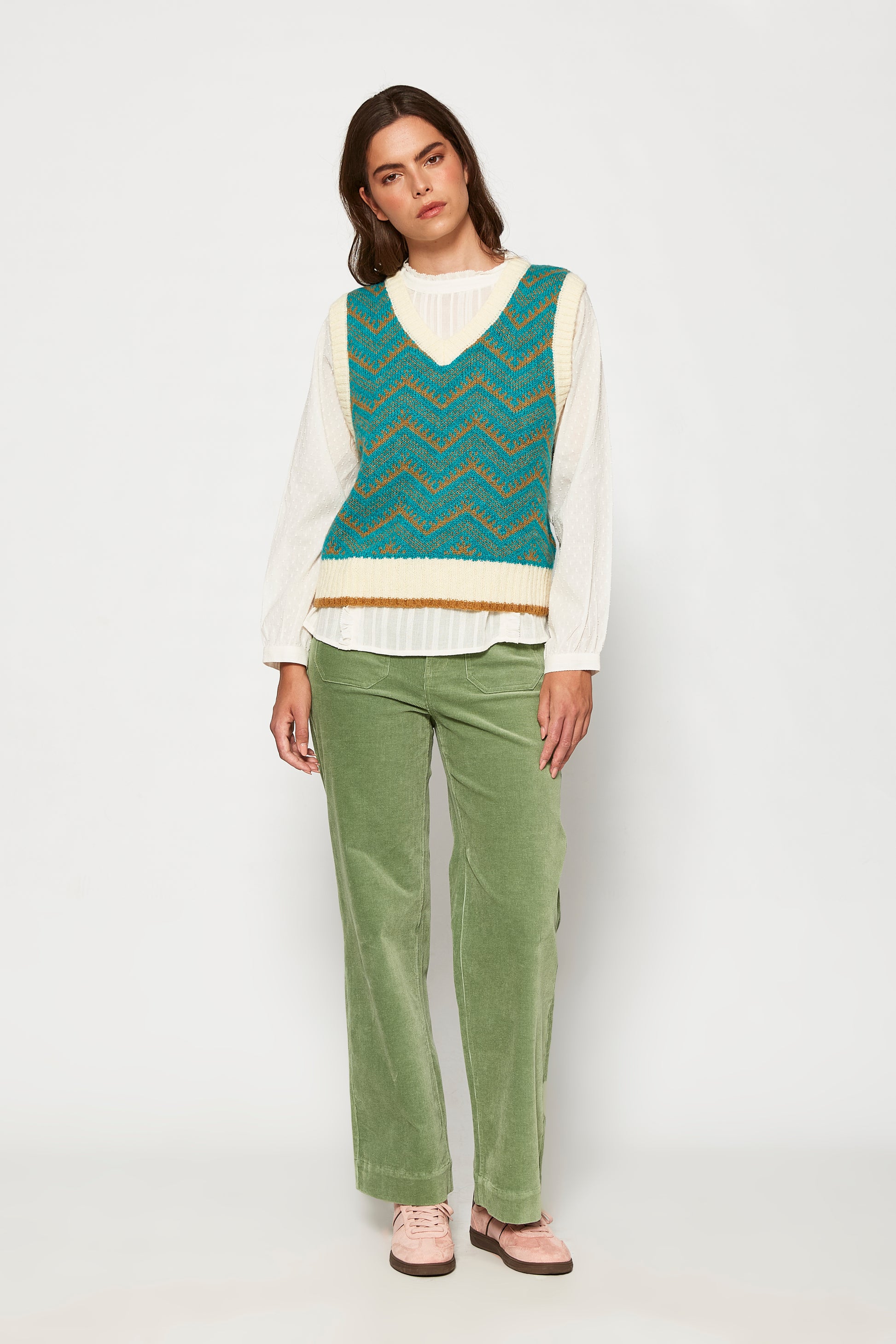 Woman wearing a teal patterned vest over a white shirt with green pants on a white background