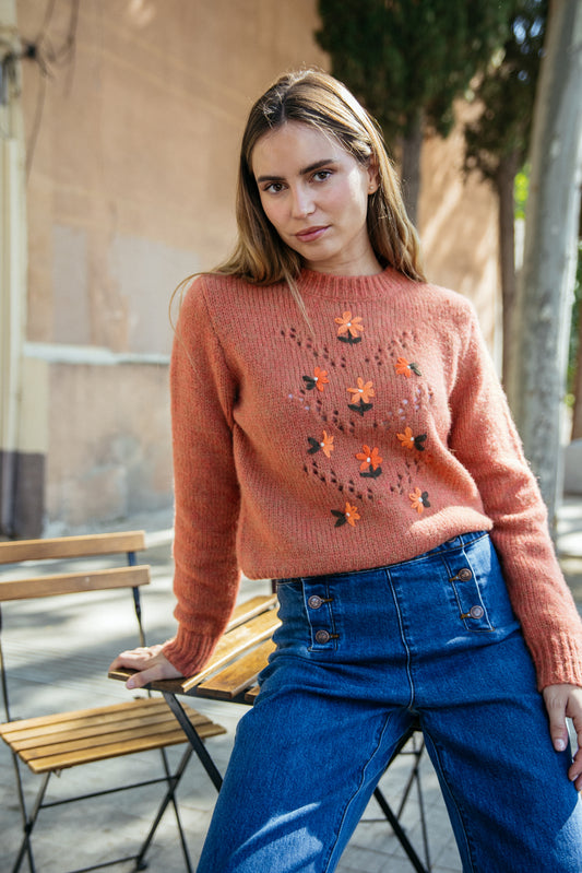 Woman wearing a pink sweater with floral patterns and blue jeans outdoors.