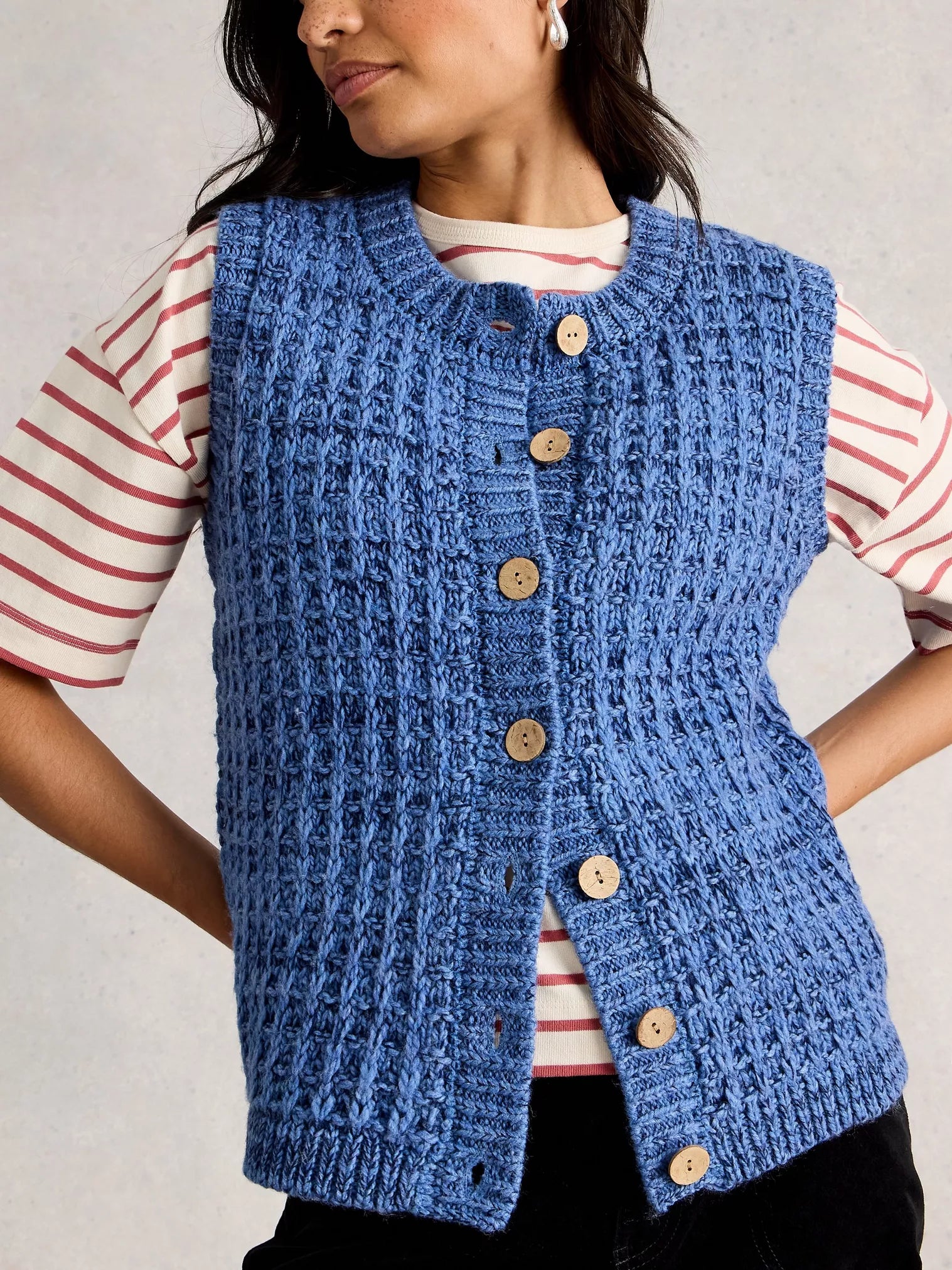 Person wearing a blue knitted vest over a striped shirt against a neutral background