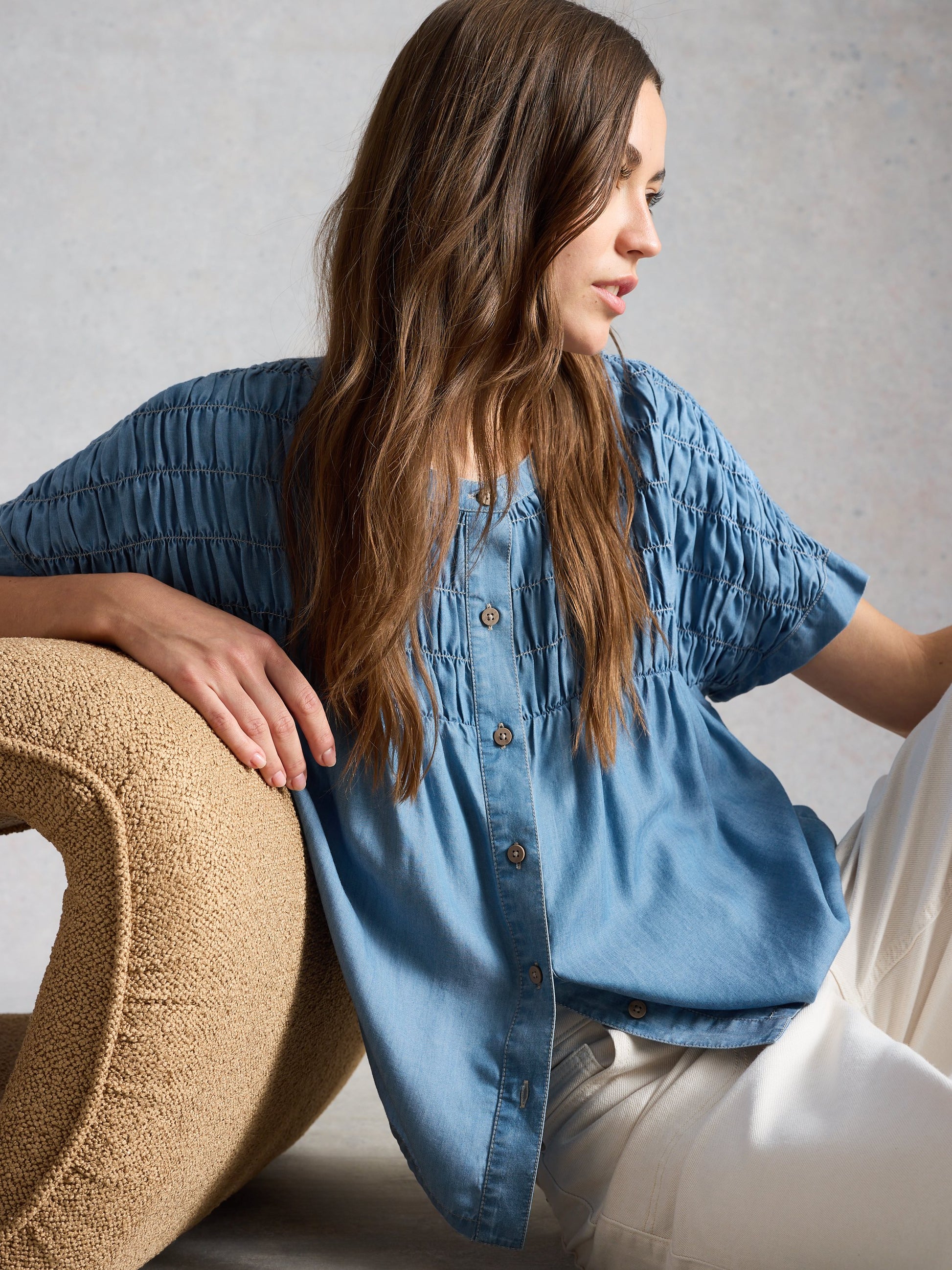 Woman wearing a blue denim shirt sitting on a textured chair against a neutral background