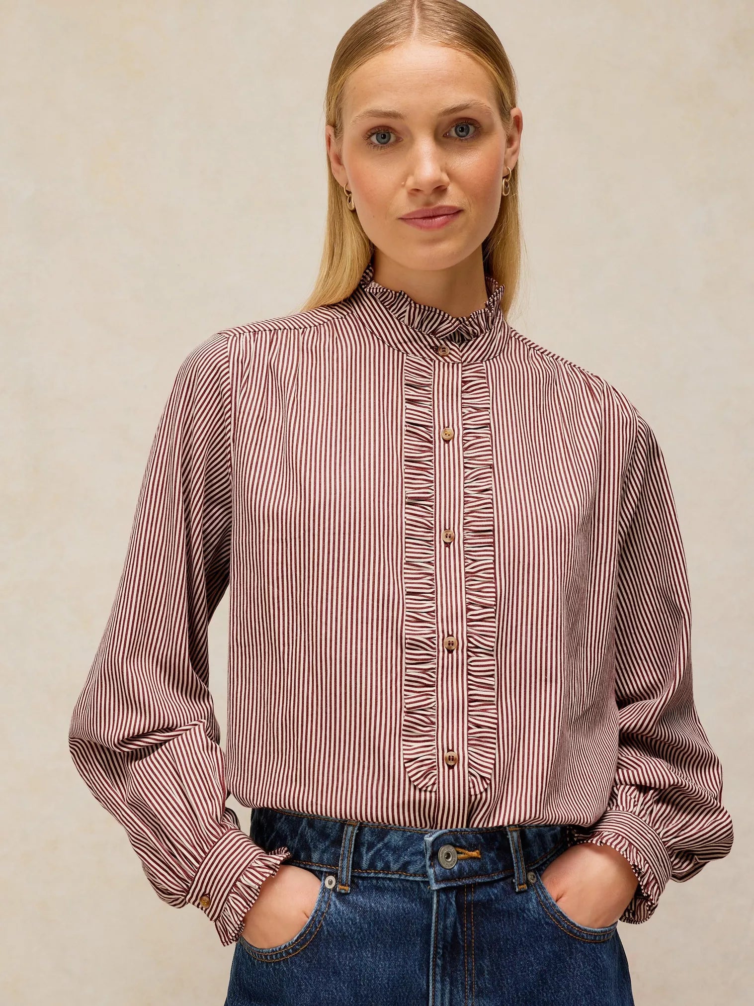 A woman wearing a long-sleeve button-front blouse with vertical stripes in dusty rose and white, paired with blue jeans, against a neutral beige background.