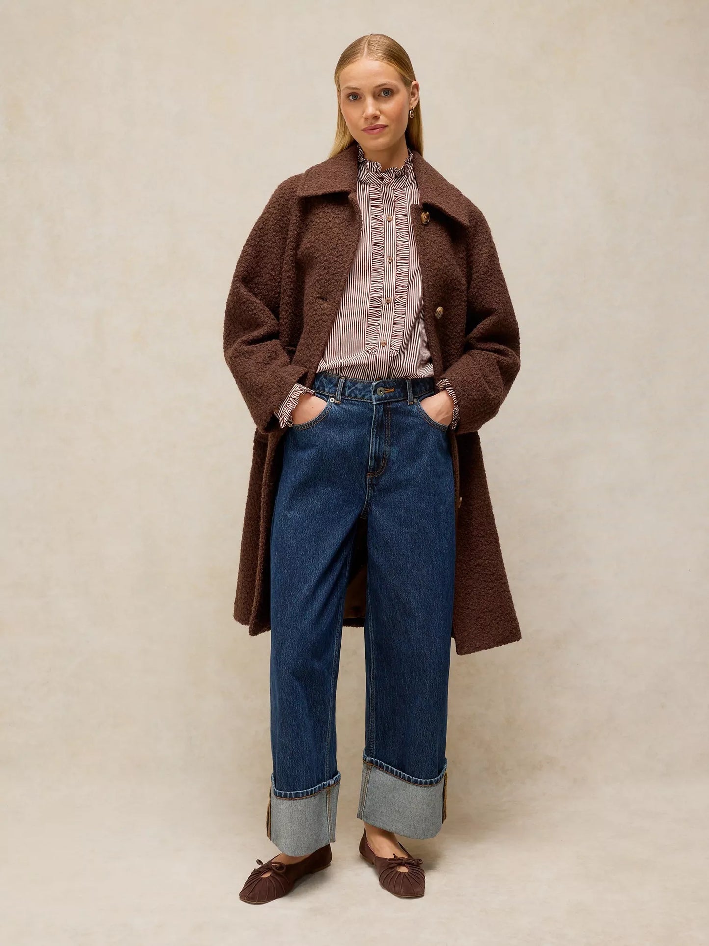 A woman stands against a neutral beige studio background wearing a dark brown textured coat over a dusty pink ruffled blouse, blue wide-leg jeans with cuffed hems, and brown loafers.