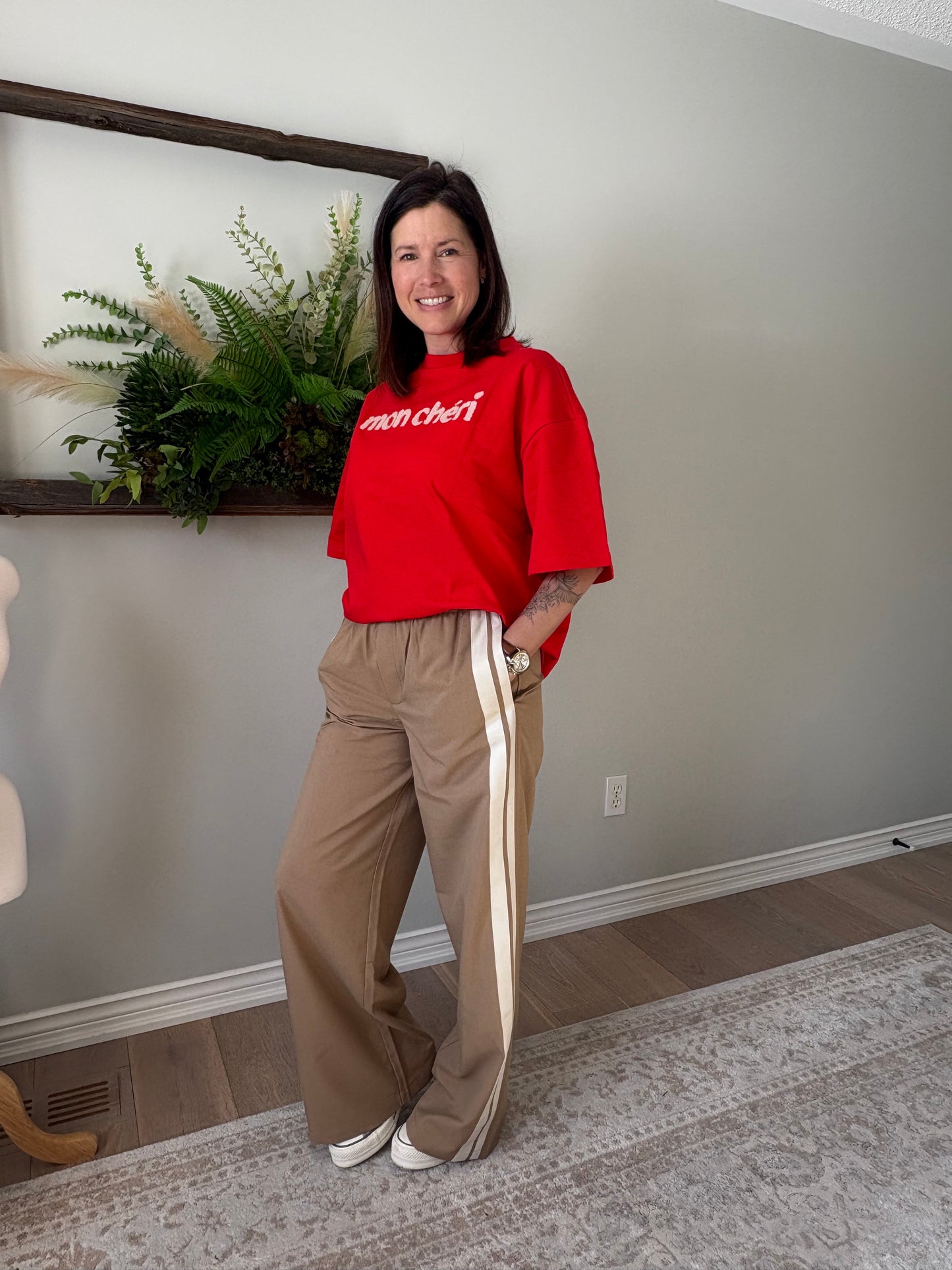 Person wearing a red sweatshirt and beige pants standing in a room with a plant and mirror.