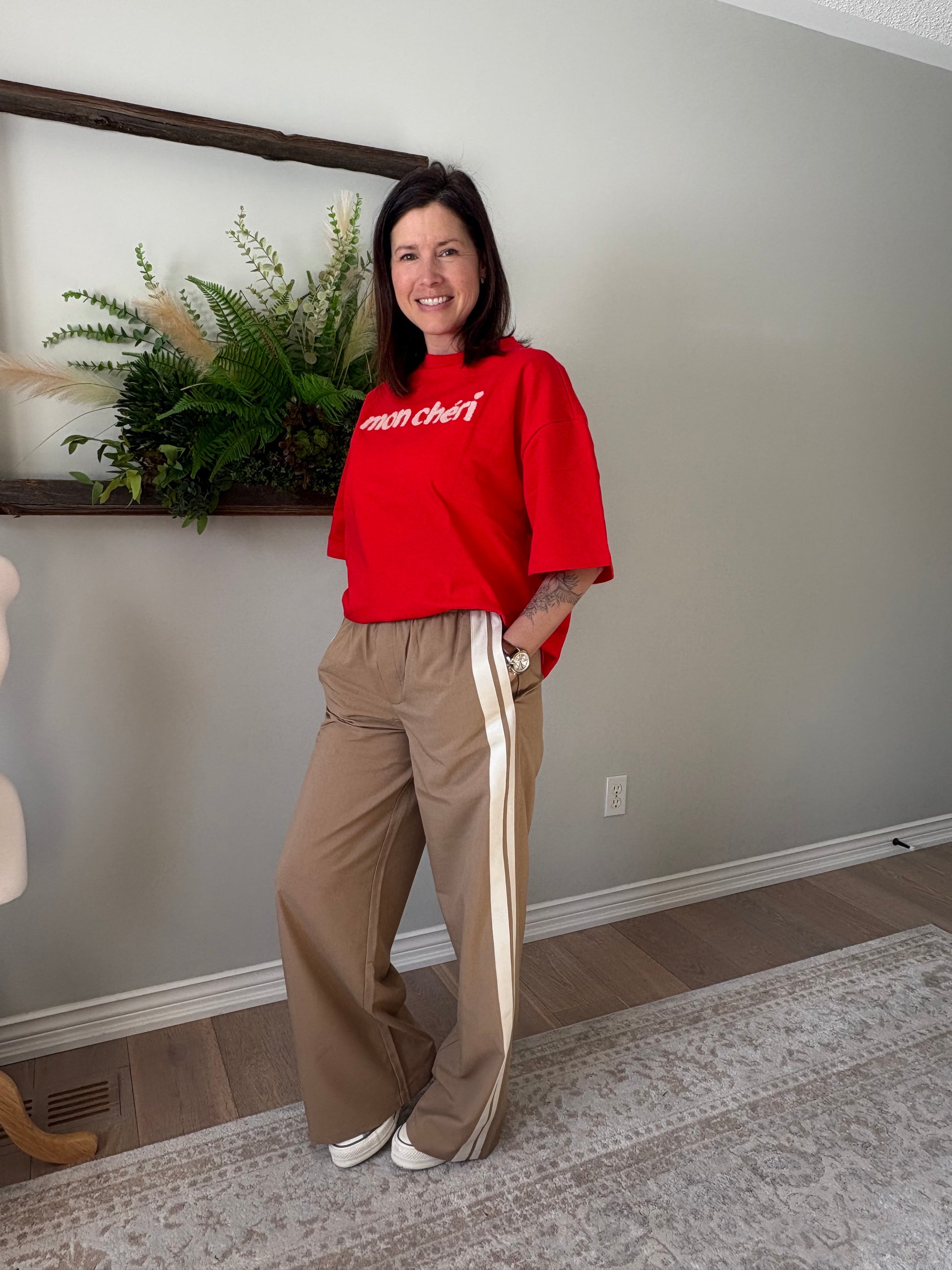 Person wearing a red sweatshirt and beige pants standing in a room with a plant and mirror.