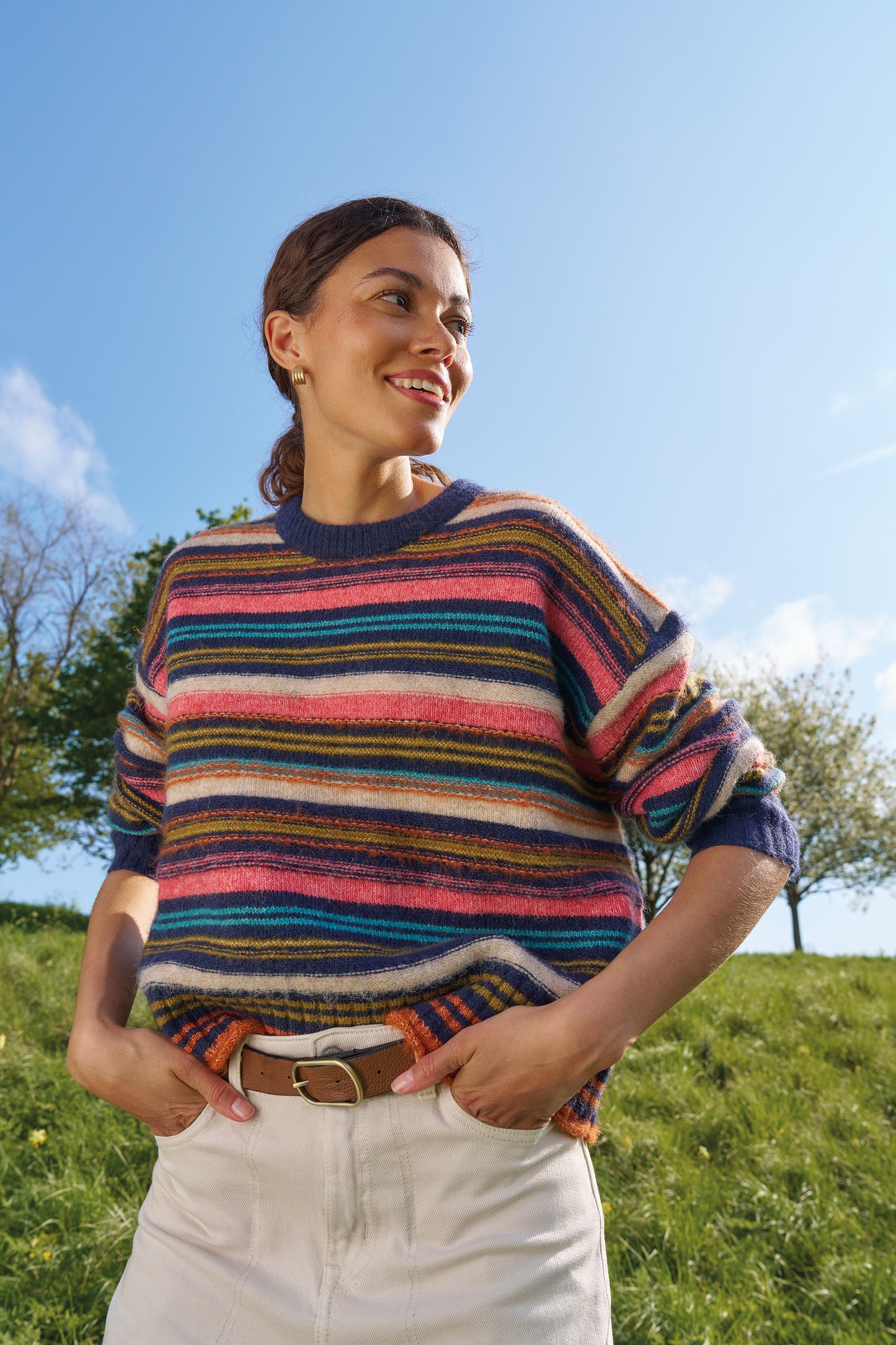 A woman wearing a colorful, fuzzy striped crewneck sweater and white pants stands outdoors on a grassy hill under a blue sky.