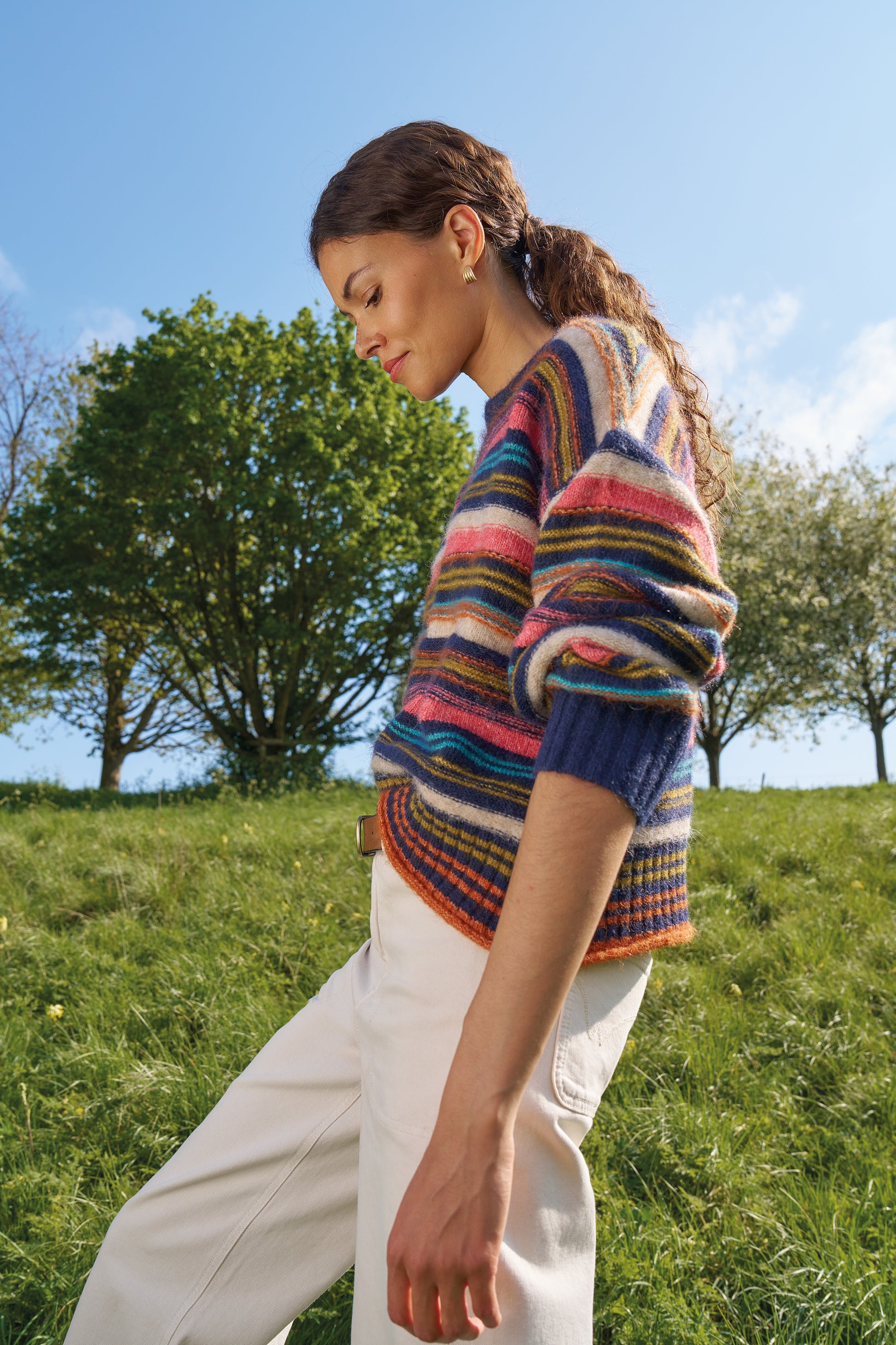 A woman wearing a colorful striped knit sweater and white trousers stands outdoors on a grassy hill under a blue sky with trees in the background.