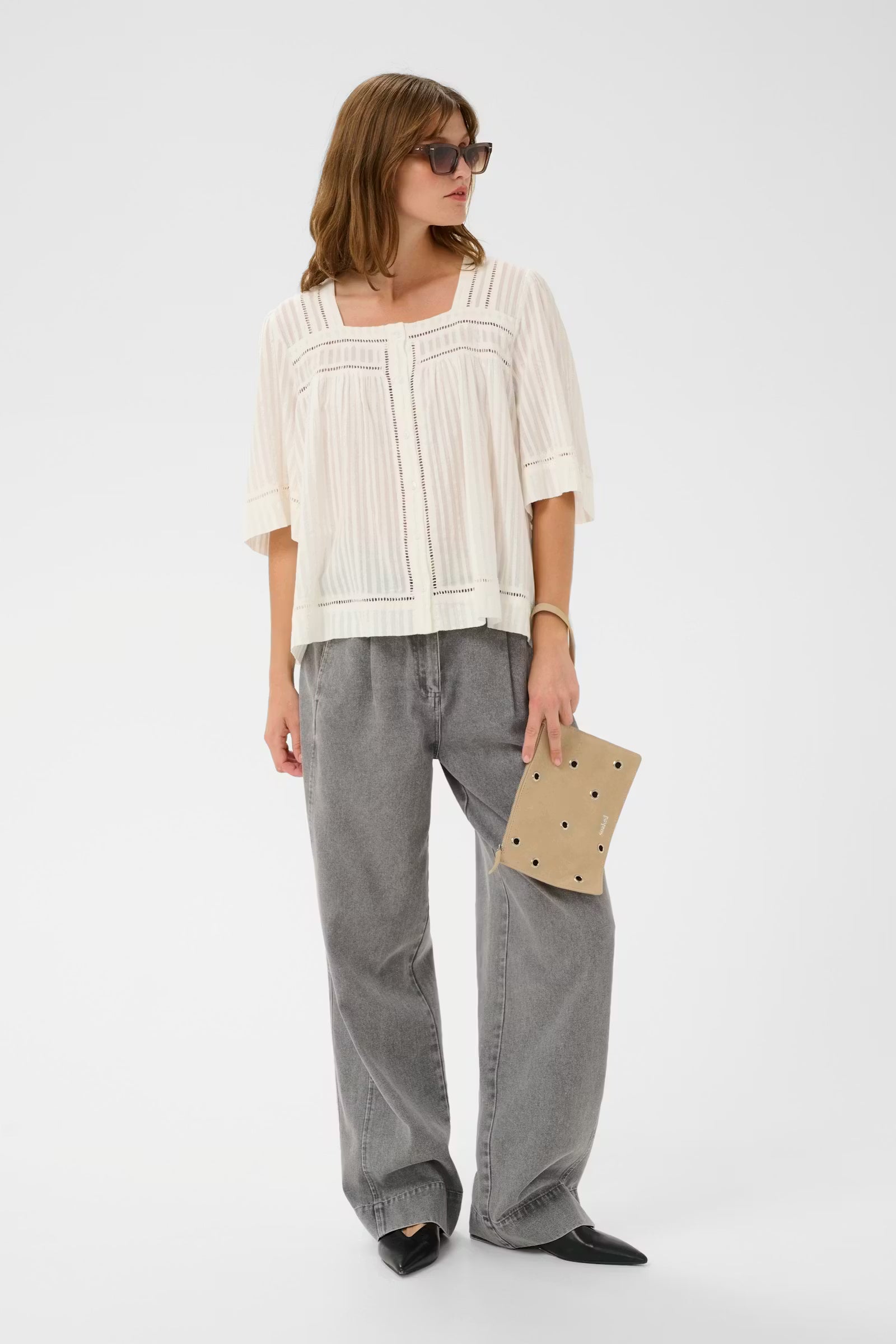 Woman wearing a white blouse and gray pants holding a beige clutch against a white background