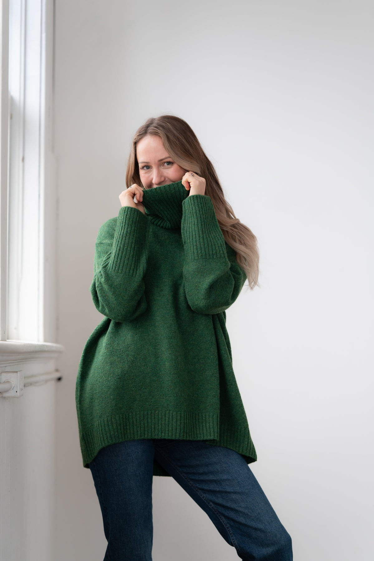 Woman wearing the Echoverde Dog Walker forest green chunky cowl neck sweater with dark jeans, pulling the cowl up over her chin