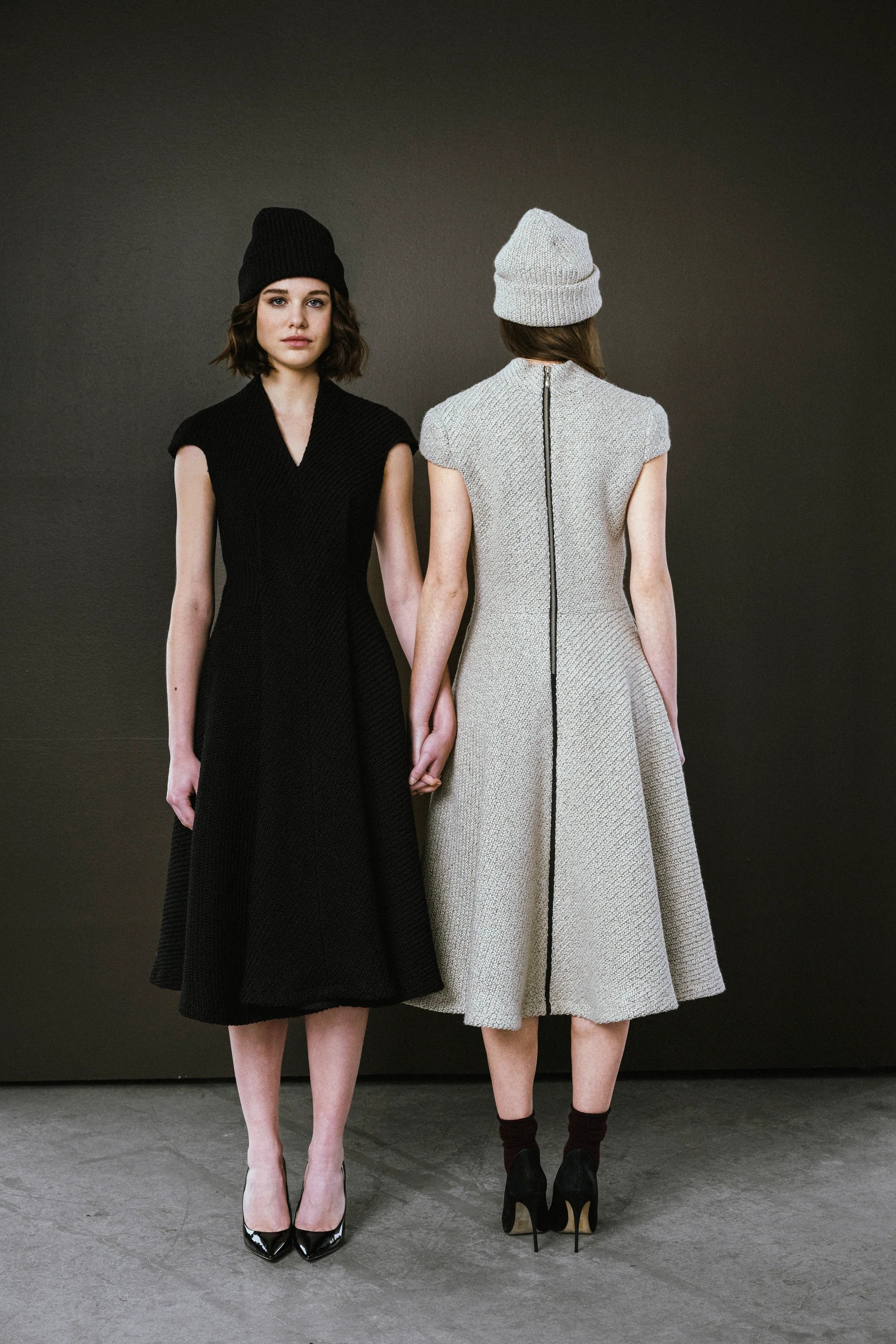 Two women holding hands, one in a black dress and black shoes, the other in a gray dress and black shoes, against a dark background.