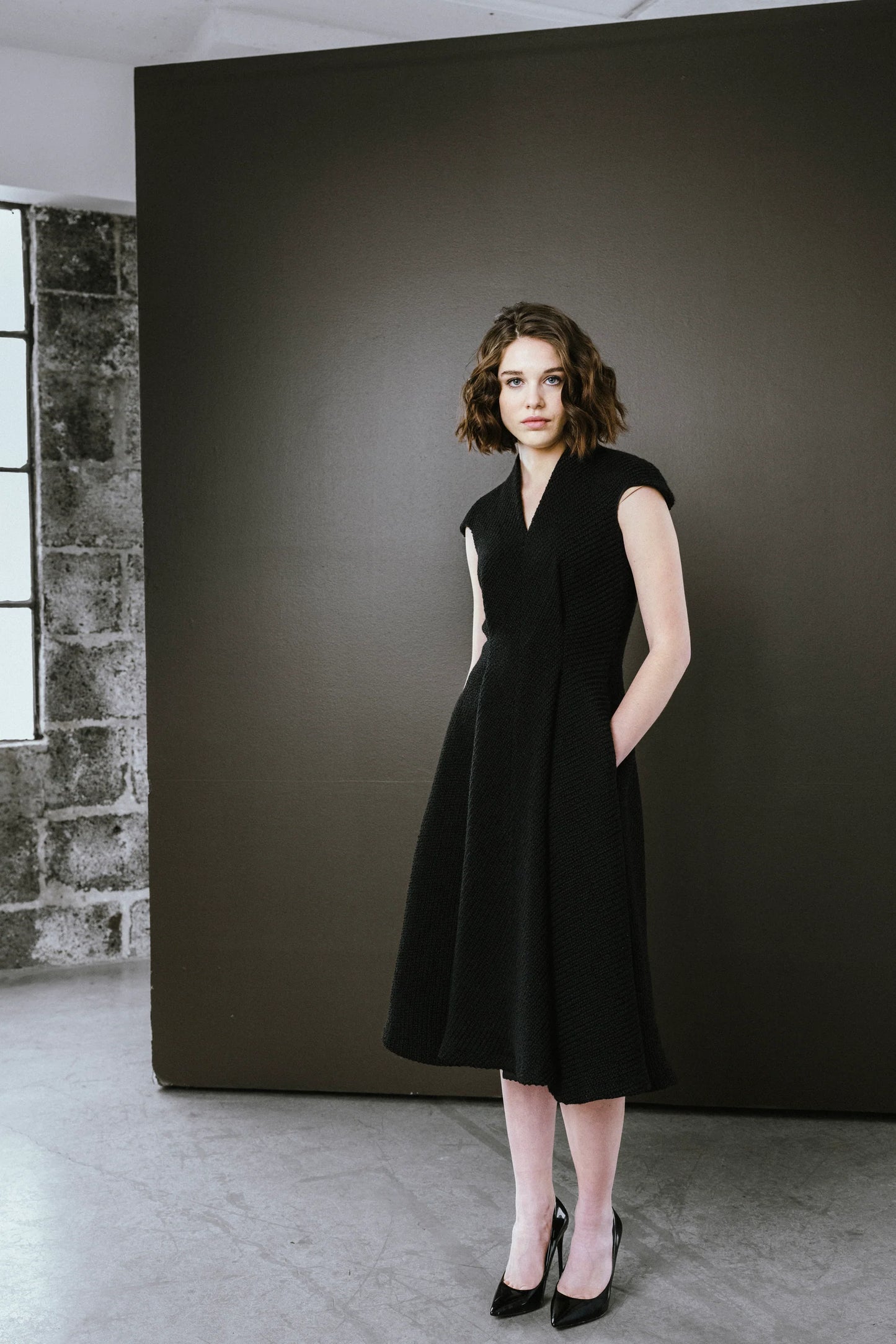 Woman in a black dress standing against a dark wall.