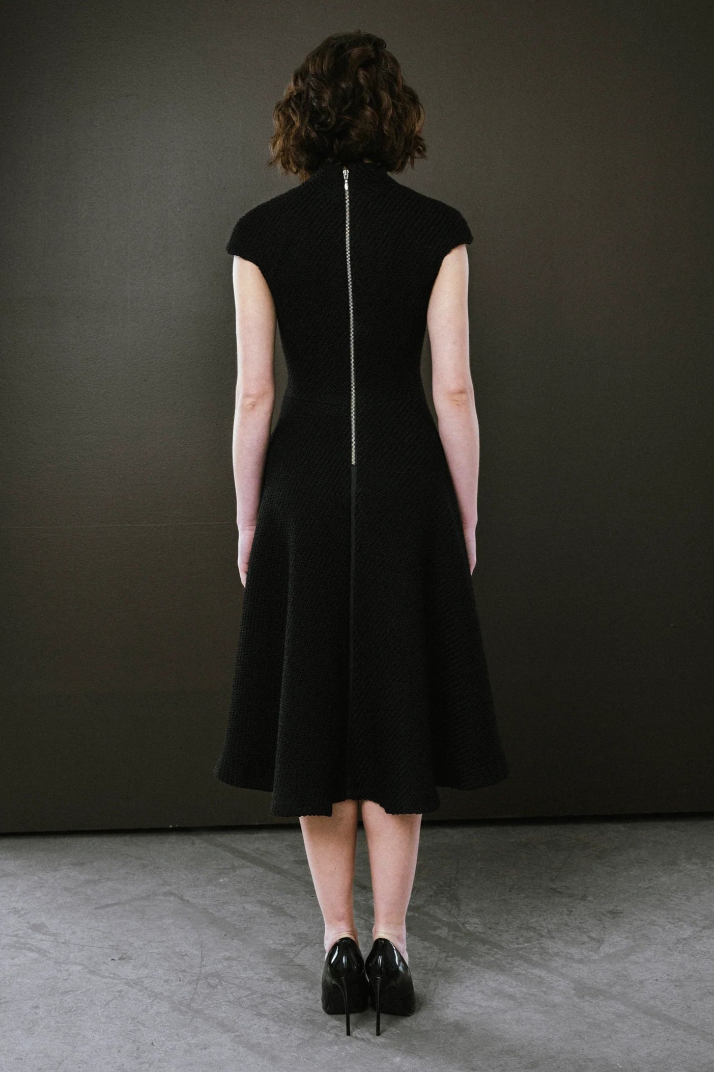 Person wearing a black sleeveless dress with a zipper back against a dark wall.