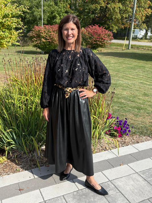 Woman in black outfit with a belt standing outdoors on a paved path with greenery.