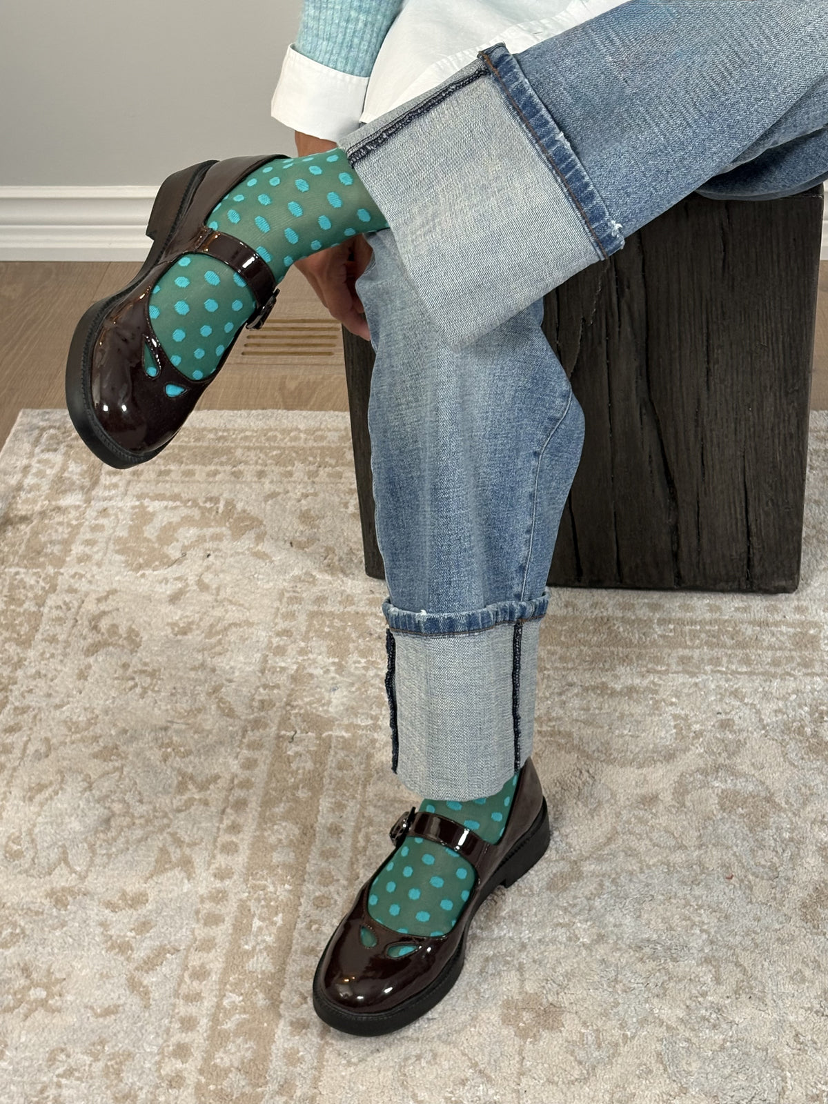 A person seated on a dark wooden stool wearing light blue cuffed jeans, shiny dark brown Mary Jane loafers, and teal socks with white polka dots in an indoor room with a patterned rug.