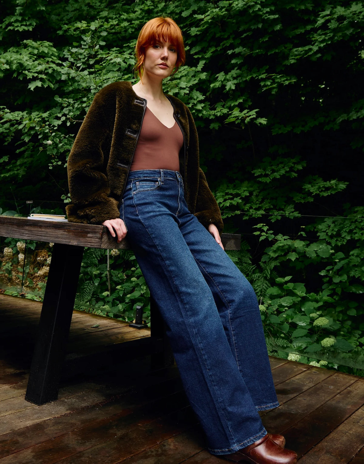 A model with red hair leans against a wooden bench in a wooded outdoor setting, wearing a dark brown fuzzy cardigan, a brown top, wide-leg blue jeans, and brown leather boots.