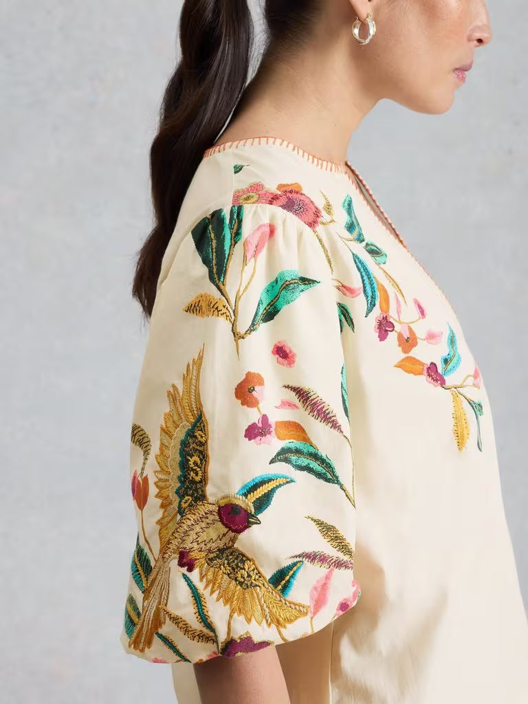 Close-up of a person wearing a floral and bird embroidered blouse against a gray background