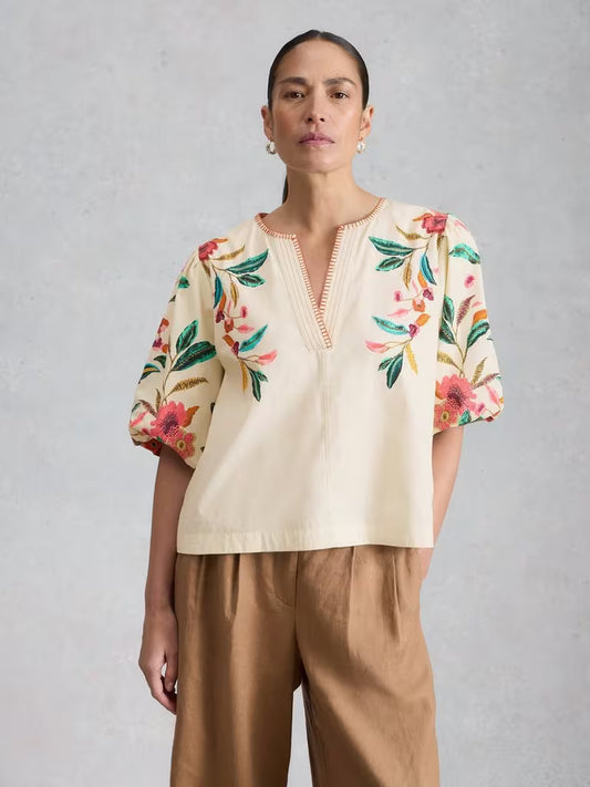 Woman wearing a cream blouse with floral embroidery and brown pants on a gray background
