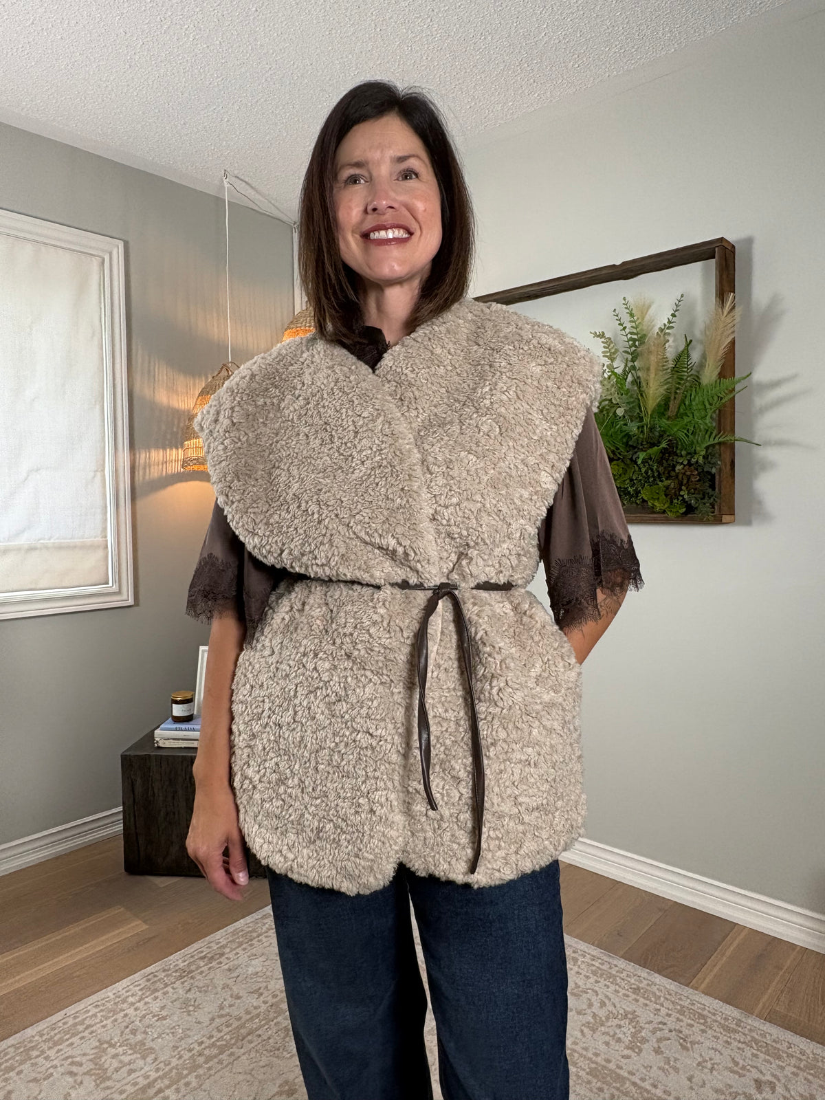 A woman wearing a chunky, textured beige sherpa vest with a dark brown tie belt over a brown top and blue jeans, standing in a home interior setting.