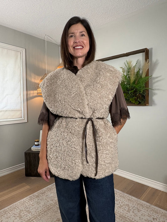 A woman wearing a chunky, textured beige sherpa vest with a dark brown tie belt over a brown top and blue jeans, standing in a home interior setting.