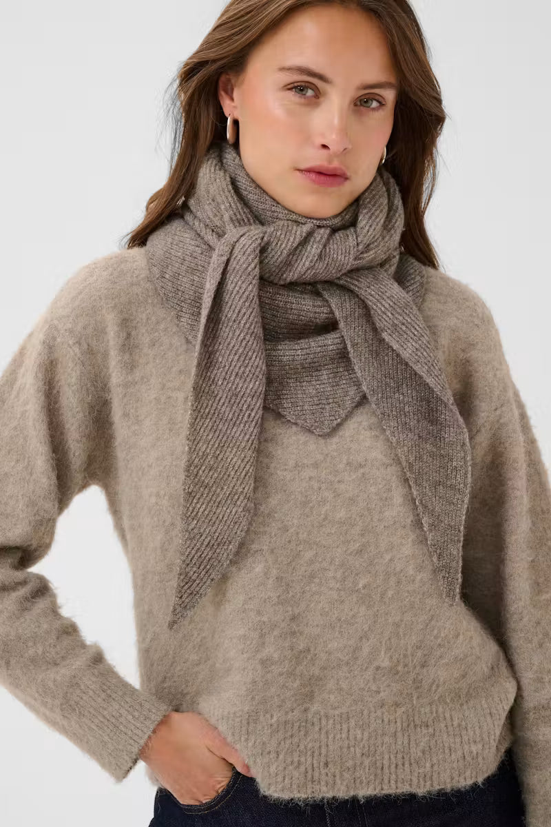 A woman wearing a soft taupe knit sweater and a matching taupe knit scarf tied around her neck, photographed against a clean light gray studio background.