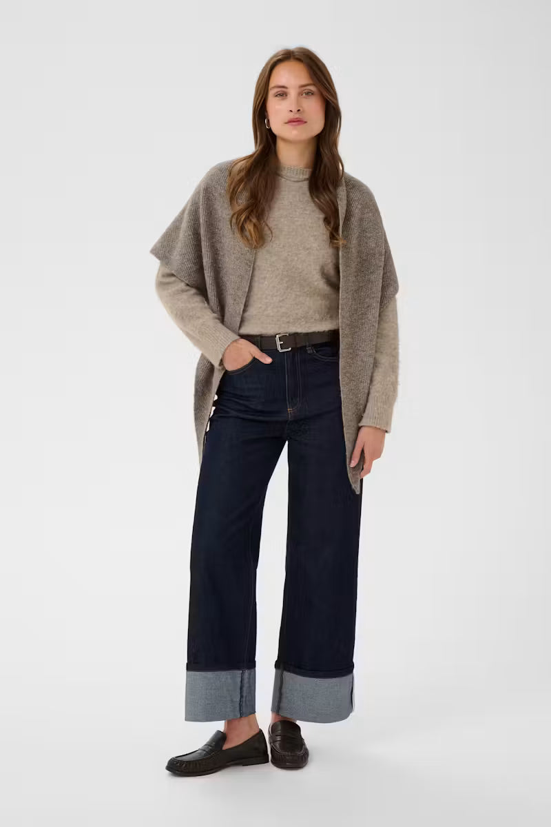 A woman stands against a light gray studio background wearing a taupe knit cardigan over a beige mock-neck top, dark blue wide-leg jeans with rolled cuffs, and dark brown loafers.