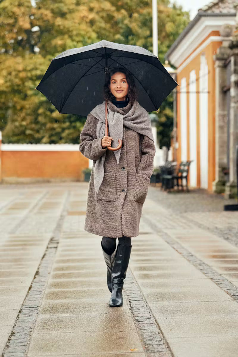 A woman walking in the rain holding a black umbrella, wearing a taupe wool coat, a light gray scarf, black tights, and black knee-high boots on a paved outdoor walkway.