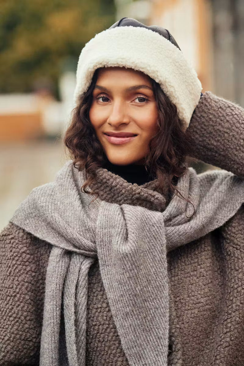 A woman wearing a cozy winter outfit featuring a cream sherpa-trimmed beanie, a chunky gray knit scarf, and a brown textured coat, photographed outdoors with a blurred background.