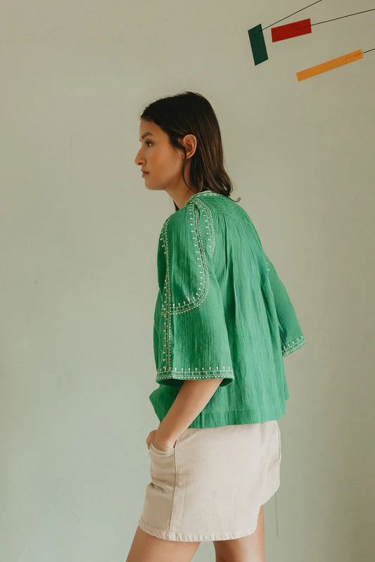 Woman wearing a green embroidered top and white shorts against a plain background
