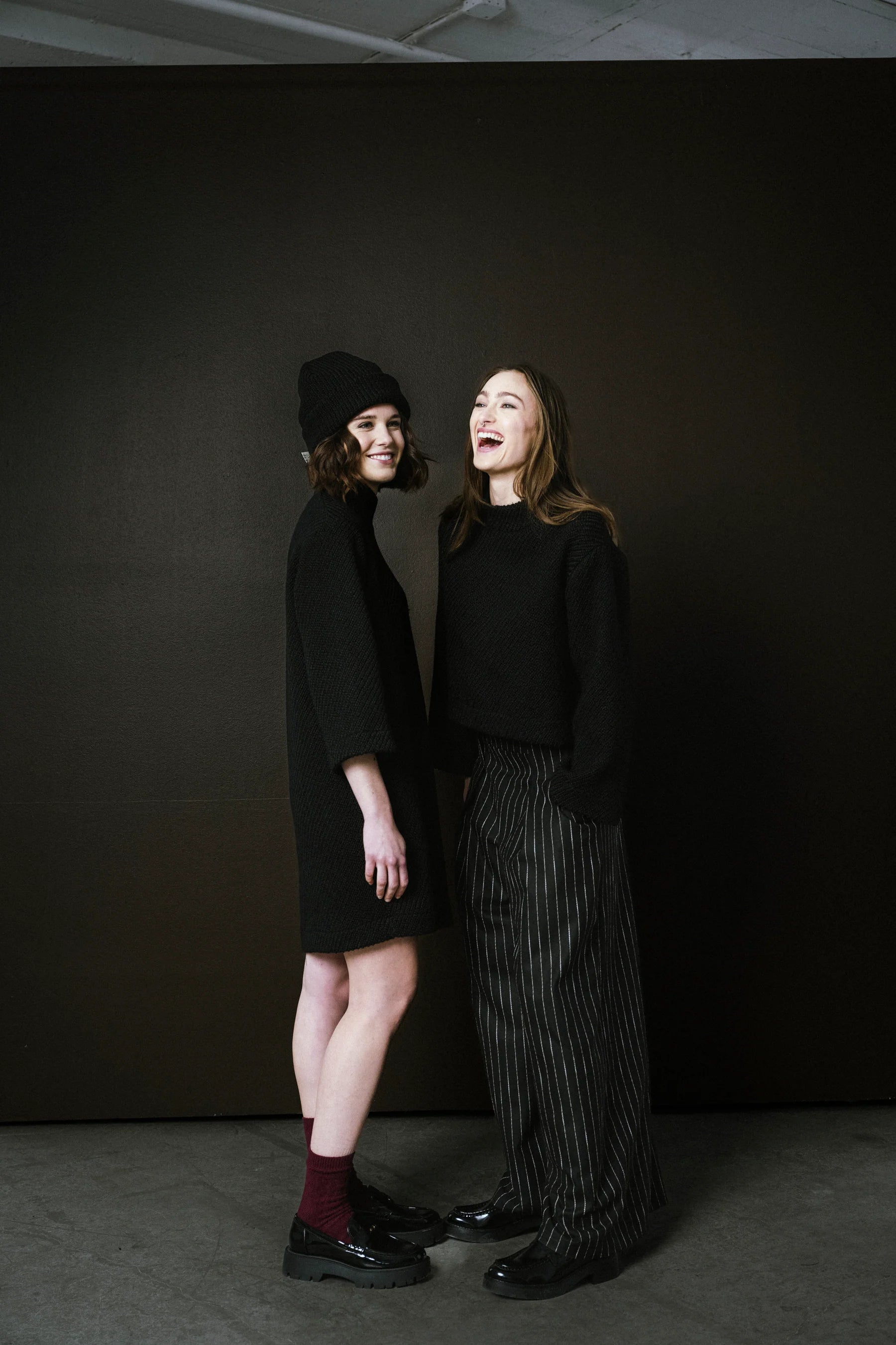 Two women standing against a dark background, one wearing a black coat and beanie, the other in a black outfit with striped pants.