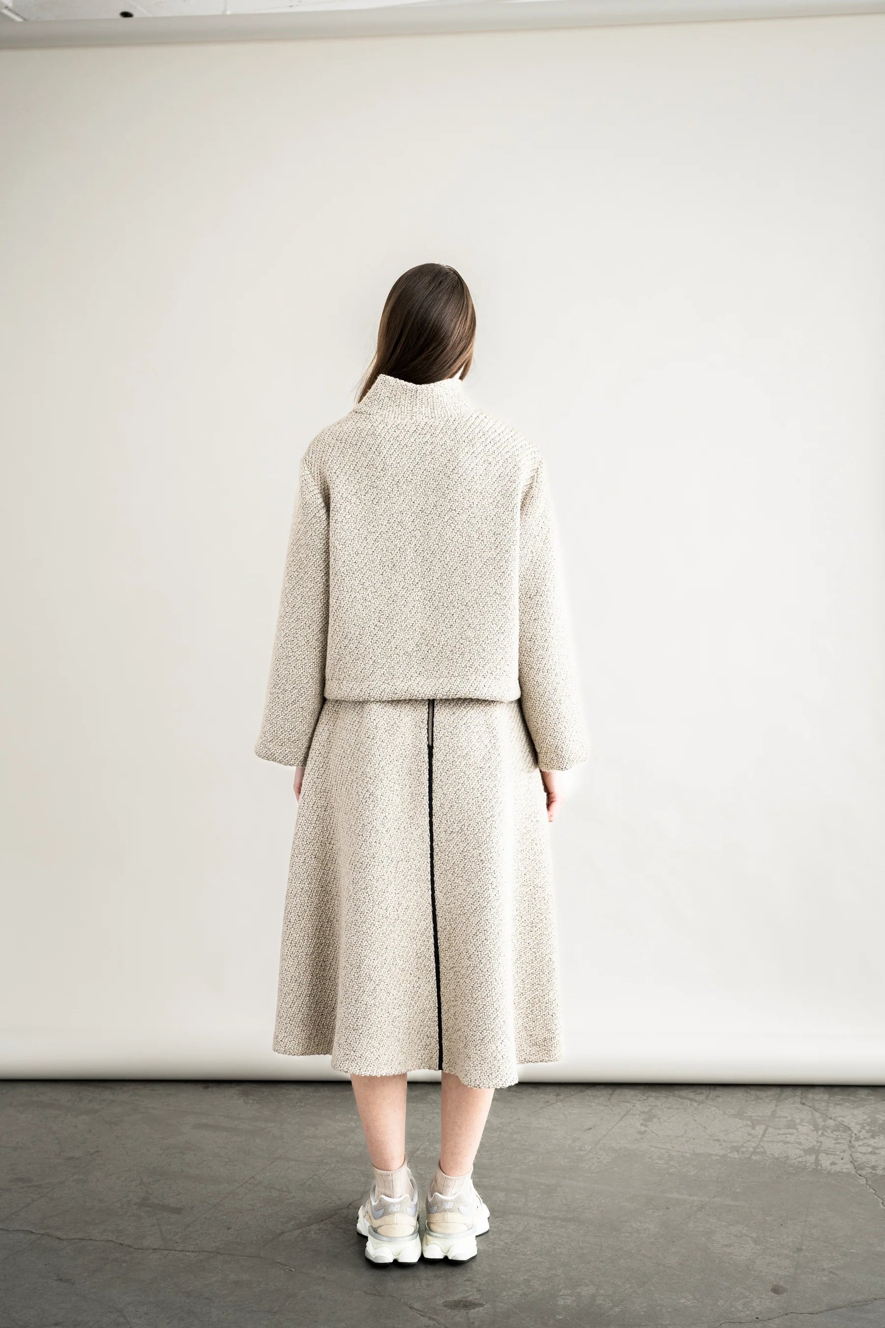 Person wearing a beige coat and skirt set against a white wall.