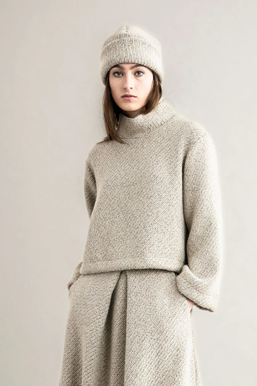 Woman wearing a beige knit sweater and hat against a plain background