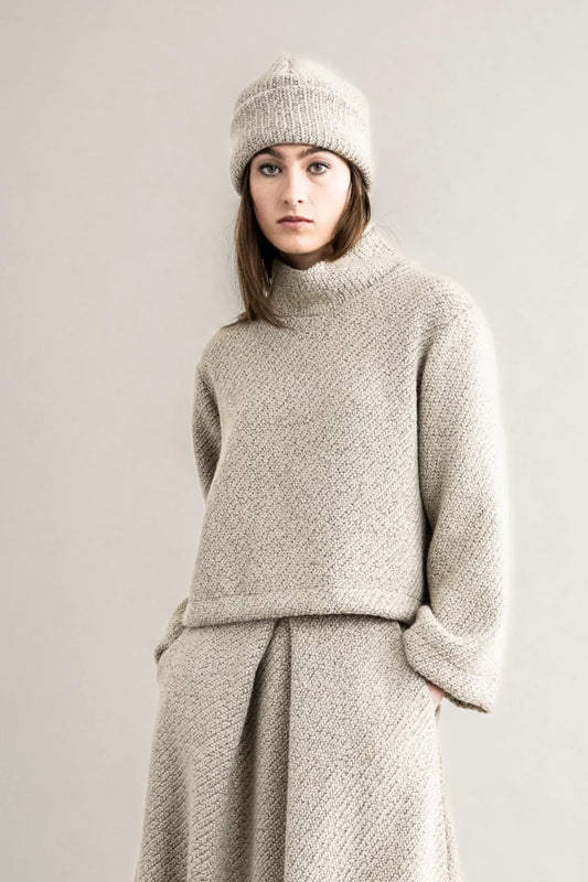 Woman wearing a beige knit sweater and hat against a plain background