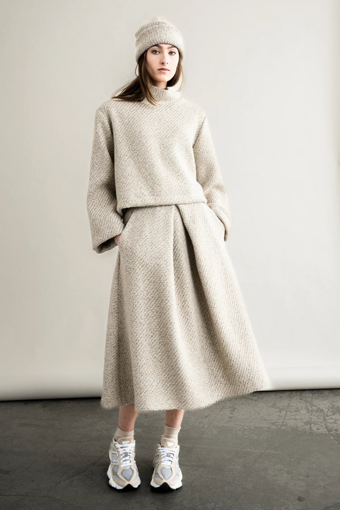 Person wearing a beige knit outfit with a skirt and white sneakers in a studio setting.