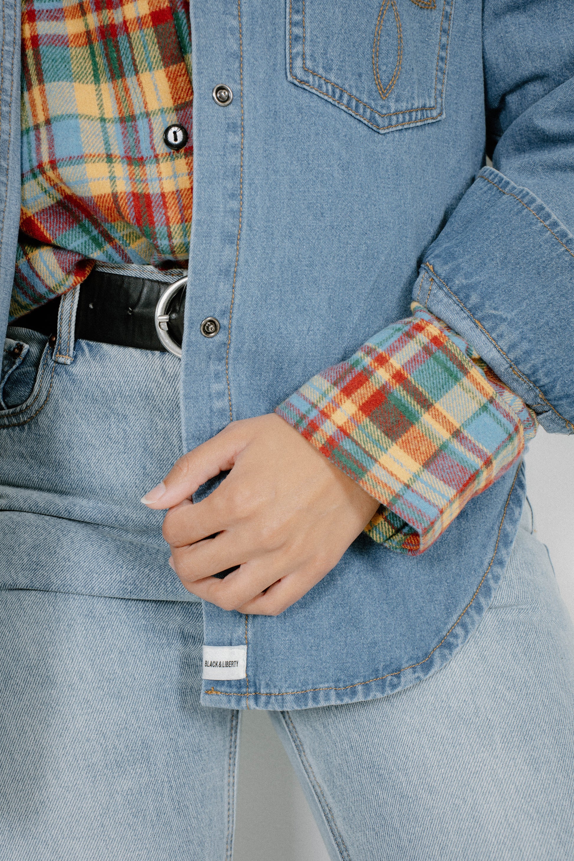 A close-up of a person wearing a light blue denim shirt-jacket over a red, yellow, and blue plaid flannel shirt, paired with light blue jeans and a black belt.